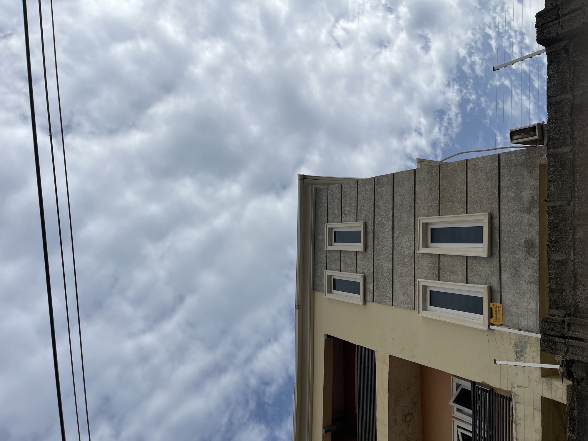 A low-angle shot shows a portion of a building and a cloudy sky. The building's facade features a combination of vertical textured panels and a cream-colored section, with small, rectangular windows. An external clothes-drying rack is fixed to the building's side, and black electrical cables stretch across the sky. The building is located in Dutsen Alhaji, Nigeria.