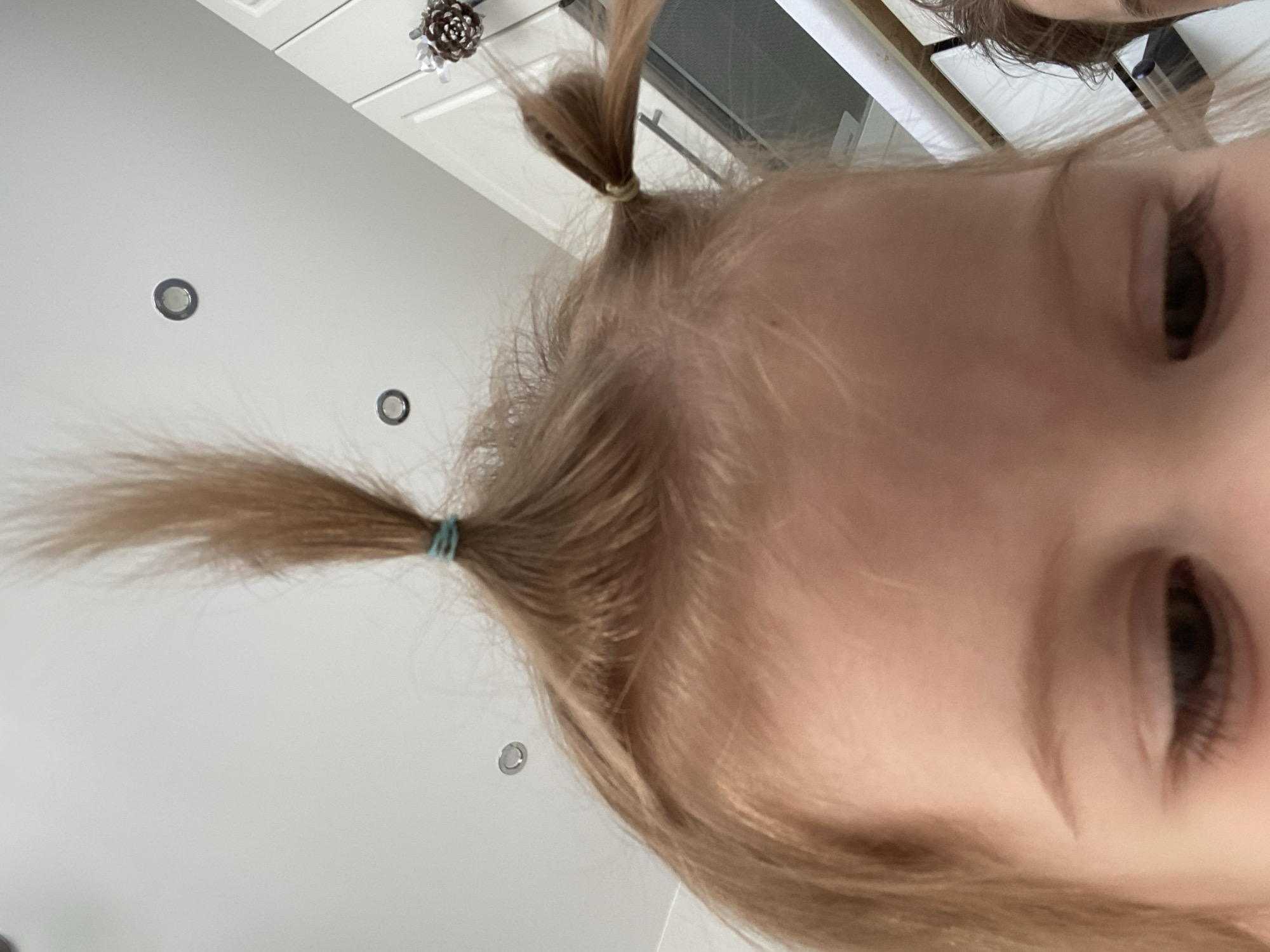 A close-up, low-angle, eye-level shot focuses on a young child's head from the forehead down to the eyes. The child's blonde hair is styled into two pigtails, one directly on top of the head and another to the right, both secured with light blue hair ties. The child's eyes are partially closed, with visible eyelashes. The background consists of a white ceiling with two circular spotlights and a hint of white cabinetry on the right, suggesting an indoor domestic environment. A small pinecone decoration is attached to the cabinetry. The overall lighting is bright and even.