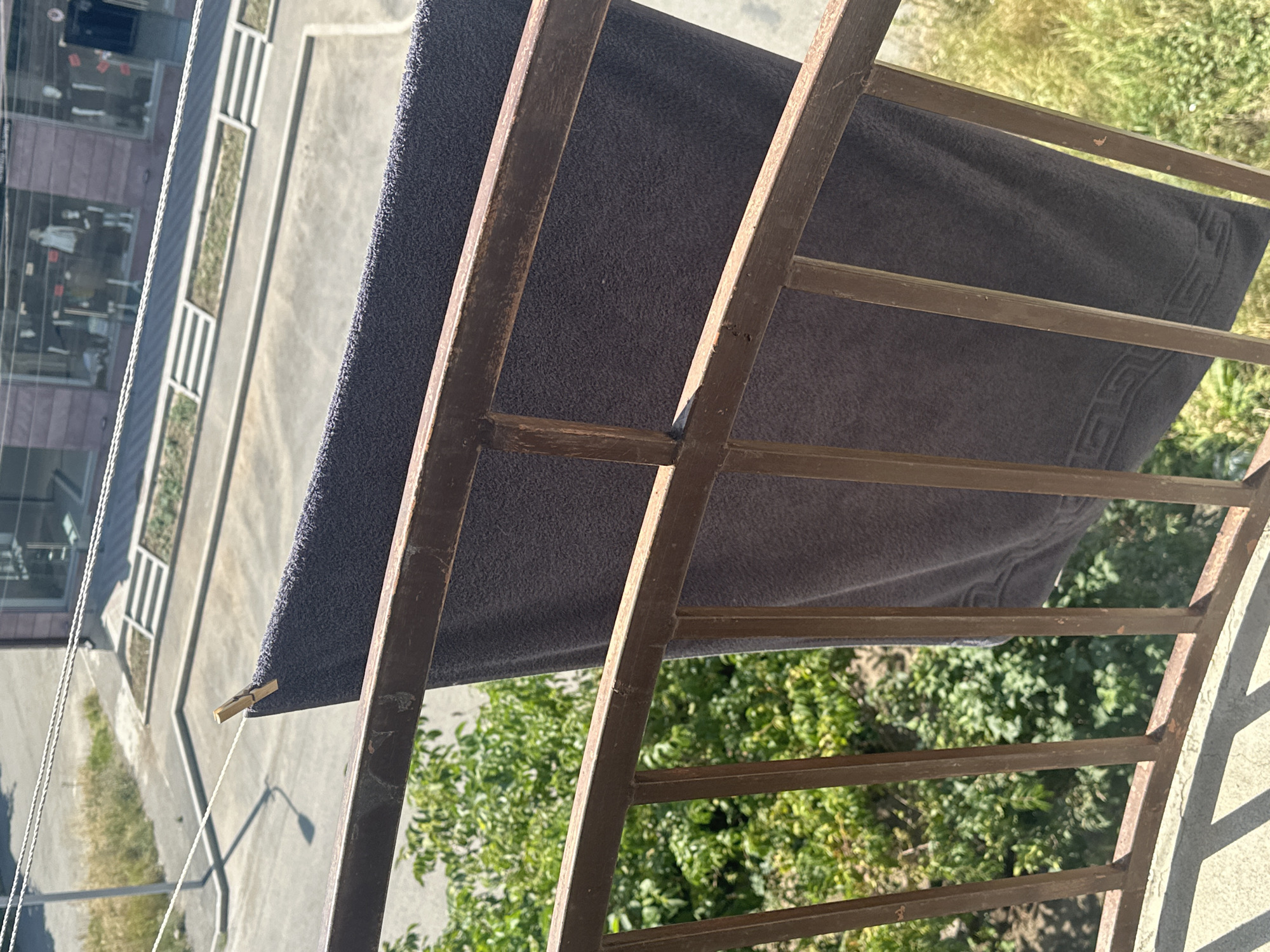 A dark gray towel is secured to a clothesline by a single wooden clothespin. The towel hangs from a rusty metal railing that appears to be part of a balcony. In the background, a street and buildings are visible, including what looks like a storefront with mannequins. Green foliage from trees and bushes is present below and to the left of the railing. The scene suggests an outdoor setting in Gyumri, Armenia, likely a residential area with commercial activity nearby.