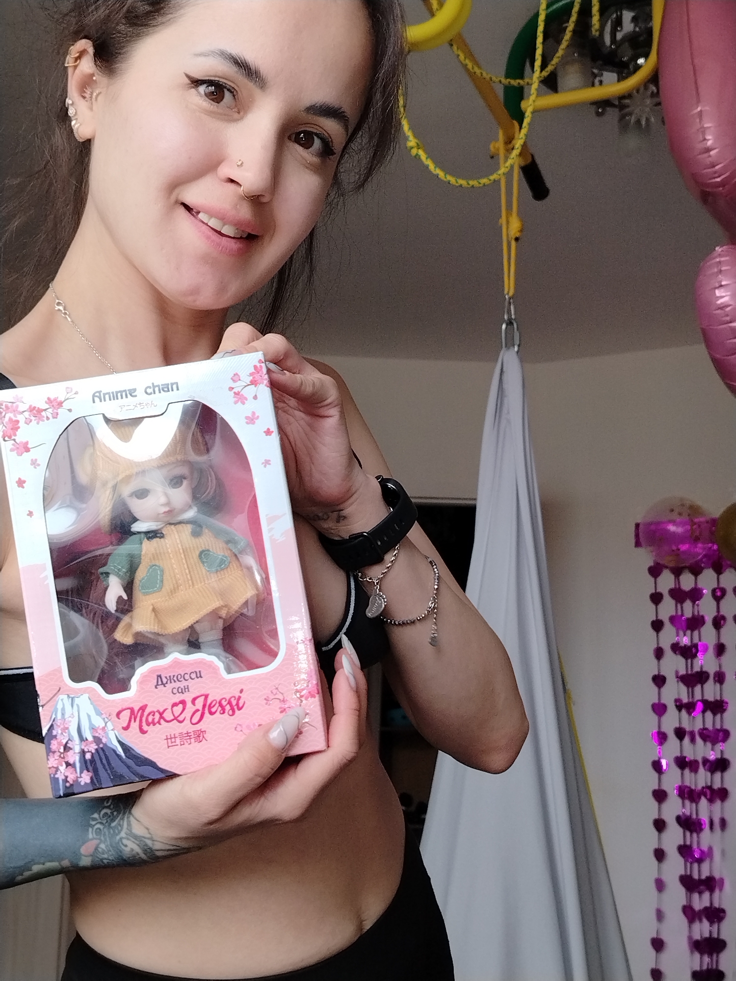 A woman with dark hair, multiple ear piercings, and a nose ring smiles as she holds up a doll in its packaging. The doll, inside a box labeled "Anime Chan" and "Max & Jessi," is dressed in a yellow dress with green hearts and a matching hat. The woman has a tattoo on her left forearm and wears a black sports bra and black bottoms. She has a black smartwatch and a charm bracelet on her left wrist. In the background, there is a yellow jungle gym, a white hammock-like fabric hanging from the ceiling, pink balloons, and a purple heart-shaped garland. The location may be inferred as Noviy, Russia, based on the Cyrillic text on the doll's packaging.