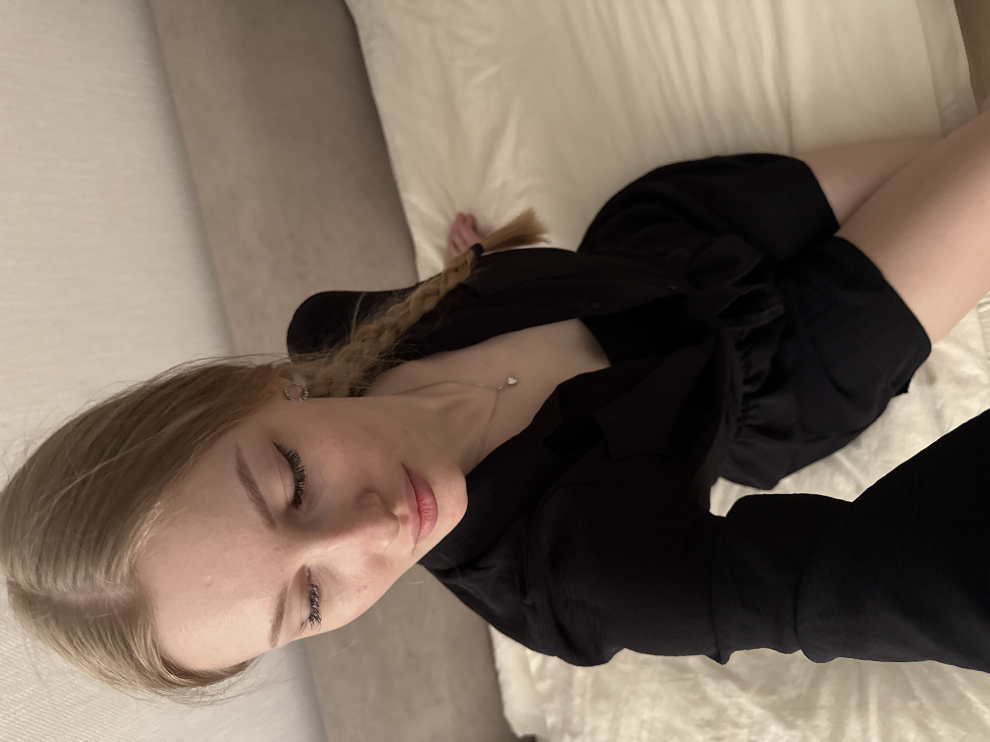 A woman lies on a white bed in a room. She has fair skin and blonde hair that is braided. She is wearing a black dress or shirt. The location is most likely indoors, perhaps in a hotel or private residence, but the city of Perm, Russia, cannot be determined from the image.