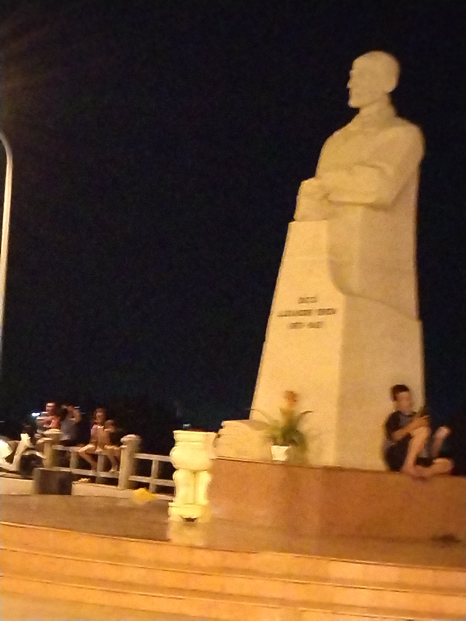 The image shows a large, light-colored monument in Nha Trang Ward, Vietnam, possibly at night. The monument is a tall structure featuring a statue of a man in a suit. The inscription on the monument is “BACSI ALEXANDRE YERSIN 1863-1943”. Several people are seen sitting on a railing and on a platform near the monument. One person is sitting barefoot on the platform of the monument, looking at a phone. A potted plant is placed on the platform. A streetlight is visible on the left side. The background is dark.