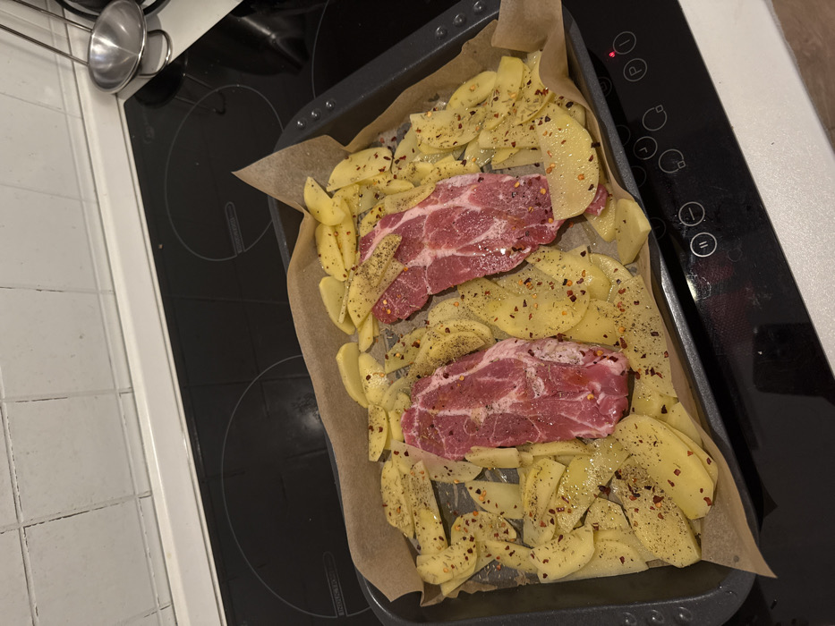 The image captures a close-up, overhead view of food preparation in a kitchen setting. The main subject is a black baking tray lined with crumpled parchment paper, holding raw ingredients ready for cooking.
Inside the tray, there are two substantial cuts of raw meat, likely pork or beef steaks, with visible marbling and a reddish-pink color. They are seasoned generously with what appears to be coarse black pepper and red pepper flakes, as well as possibly dried herbs, which are also scattered over the other ingredients.
Surrounding the meat are numerous slices or small wedges of raw, peeled potatoes. These potatoes are also seasoned with black pepper, red pepper flakes, and possibly dried herbs, and they appear to be lightly coated in oil or a similar liquid, giving them a slight sheen. The arrangement suggests they are prepared for roasting or baking.
The baking tray rests on a black induction or ceramic stovetop, which has several white control symbols and lines, including what looks like a 'P' and a red indicator light that is illuminated, possibly indicating a heating zone is active or a power status. The stovetop surface is glossy and shows some reflections.
To the left of the stovetop, part of a white tiled kitchen wall or backsplash with dark grout lines is visible. In the upper-left corner, a small, polished metal measuring cup or pot with a handle is seen hanging from a rail, partially obscured. To the right of the stovetop, a clean, white countertop or appliance surface is visible.
The overall impression is a domestic kitchen scene, showing the initial stages of preparing a meal before it is cooked. The lighting suggests an indoor setting, possibly daylight or artificial kitchen lighting.