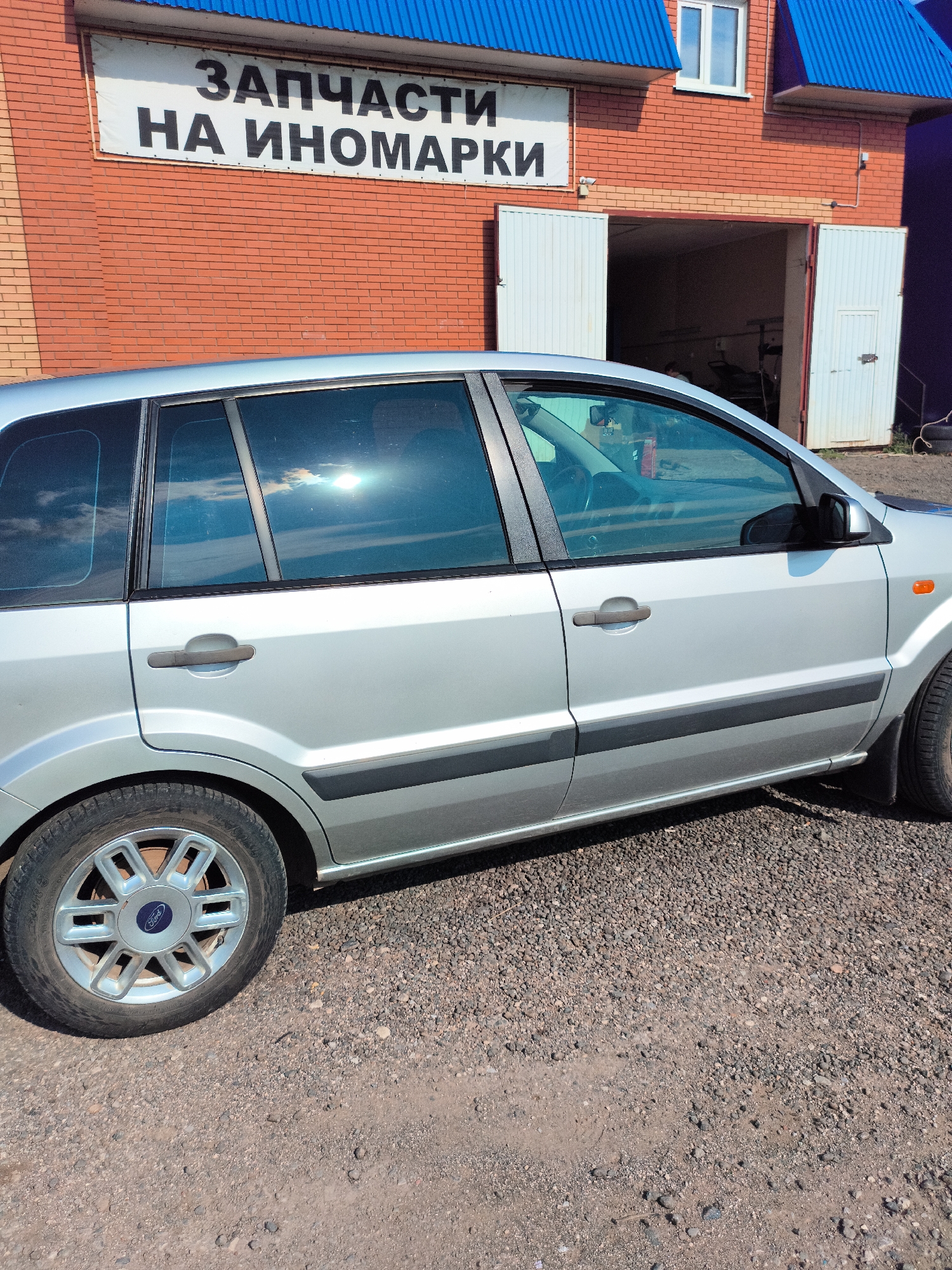 A silver Ford Fusion is parked on a gravel lot in front of a brick building. The building has a blue roof and a sign that reads "ЗАПЧАСТИ НА ИНОМАРКИ" which translates to "SPARE PARTS FOR FOREIGN CARS". The building has two large white garage doors, one of which is open, revealing the interior of a workshop. The car is facing to the right, and its front end is not fully visible. The sun is shining brightly, reflecting off the car's windows. The environment appears to be a commercial area, possibly an auto repair shop.