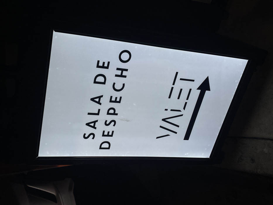 A tilted, illuminated sign with a dark frame is visible against a dark background. The sign displays the text "SALA DE DESPECHO" vertically on the left. To the right, "VALET" is written vertically, with an arrow pointing upwards alongside it. All text and the arrow are in black. The sign's background is white and shows minor blemishes. The lower left corner shows a partially visible white object.