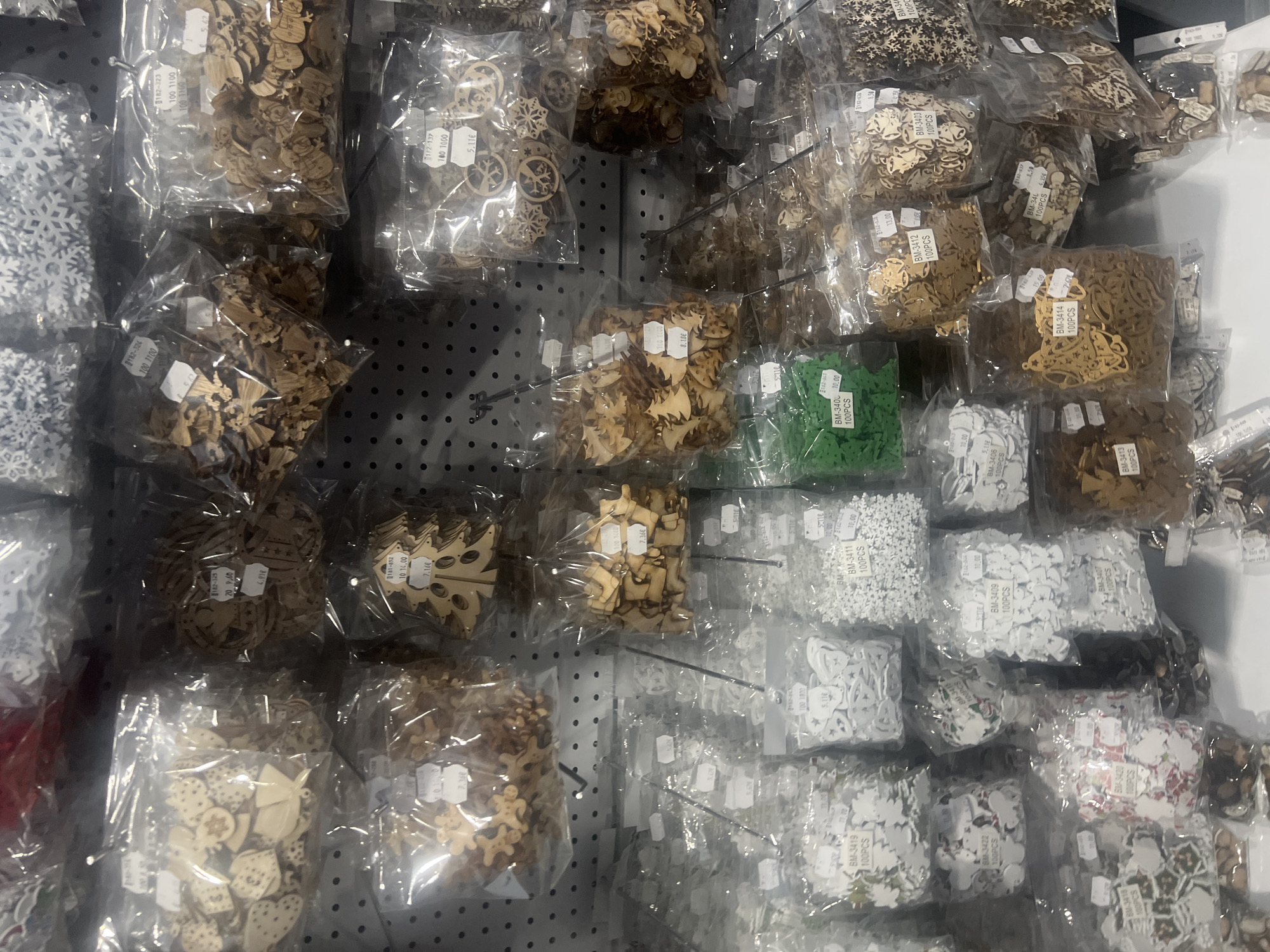 The image shows a display of small craft embellishments packaged in clear plastic bags, arranged on a perforated wall panel. The embellishments appear to be made of wood and plastic and are various shapes, including snowflakes, trees, gingerbread people, bells, and other decorative items. Several bags of the items are priced, with labels indicating "100PCS" and prices varying from 2.91€ to 20.96€. Some labels feature product codes like "BM-3419," "BM-3411," and "182-325." The scene appears to be inside a retail store. The image has been taken with a mobile phone.