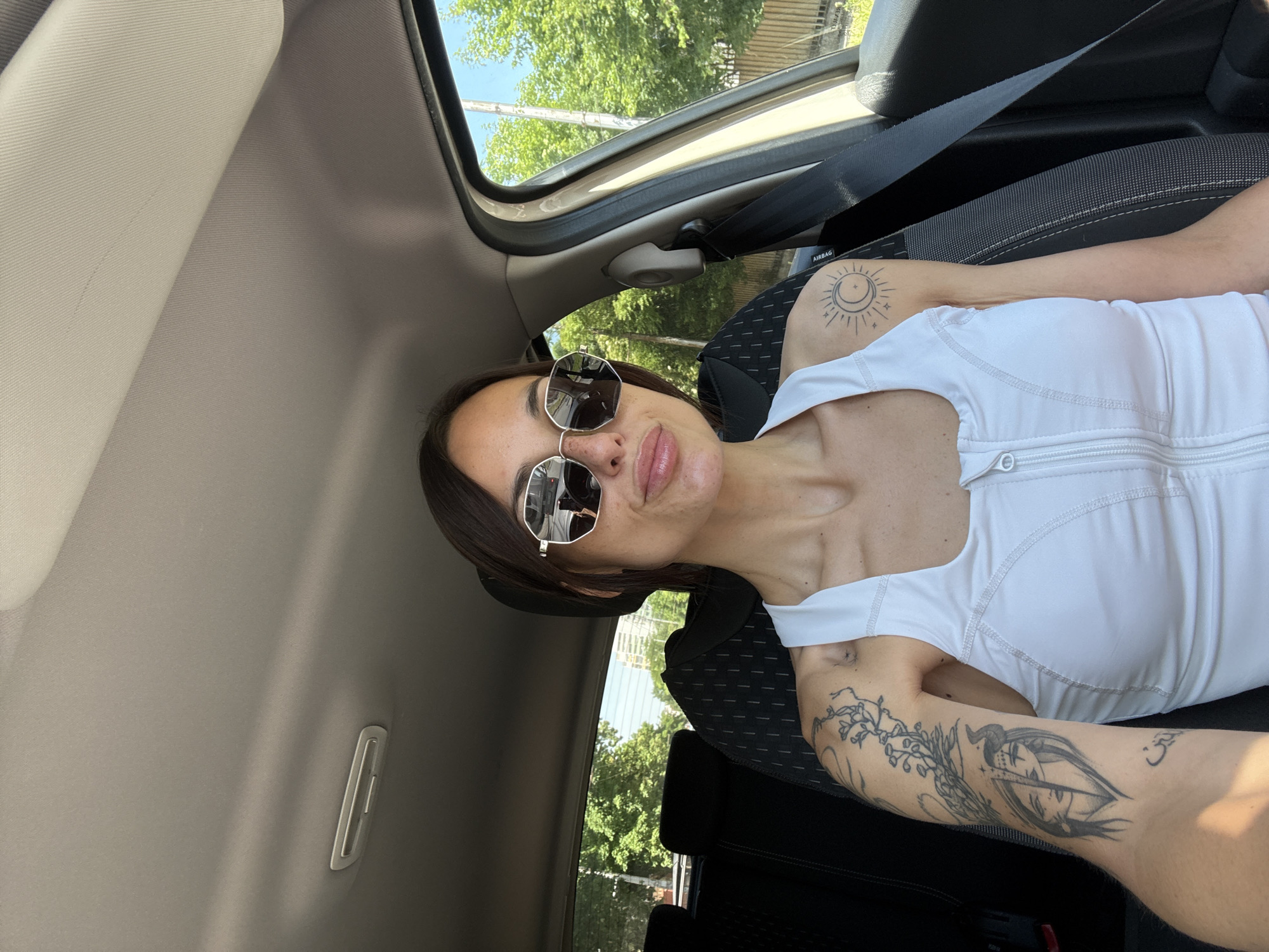 A woman with shoulder-length dark hair and wearing sunglasses and a white tank top is sitting in the driver's seat of a car. She has tattoos on both arms; the right arm features a sun and moon design, while the left arm has more complex, illustrative tattoos. The car's interior is visible, including the grey headliner, the driver's side window, the black seat, and a seatbelt across her chest. Outside the car window, green trees and a glimpse of the sky are visible.
The specific location in Pleven, Bulgaria, cannot be determined from the image.