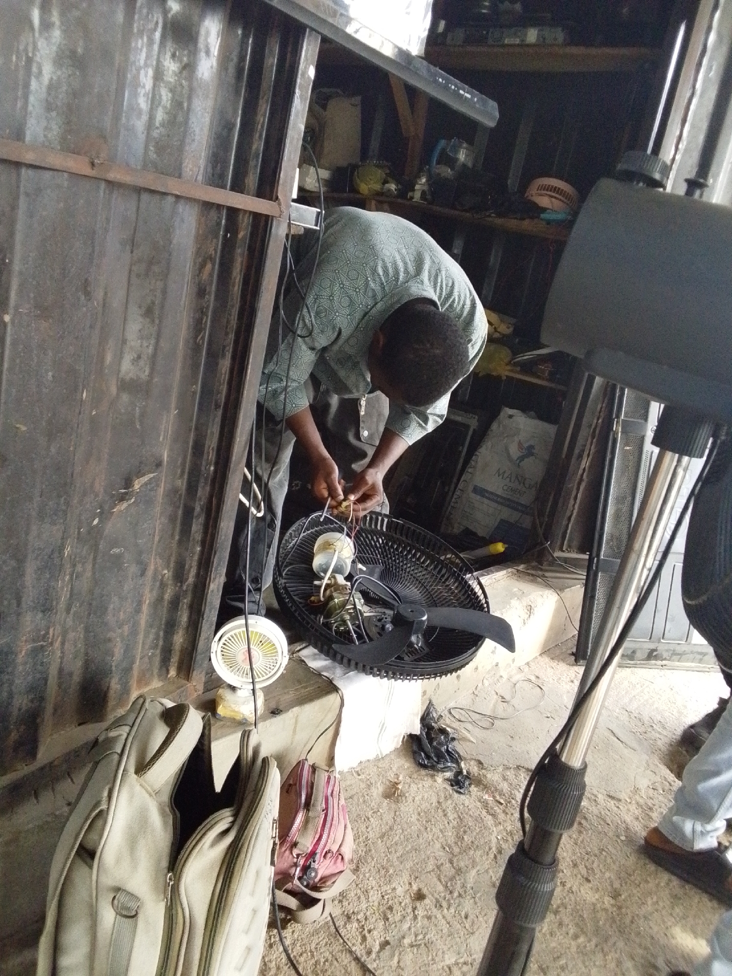 In Lokoja, Nigeria, a man is repairing a fan inside a corrugated metal structure with an open doorway. The man wears a patterned shirt and dark pants, focused on the fan in front of him. The fan is black with some parts disassembled. Nearby is a smaller white fan. A tan bag and a pink backpack sit on the floor next to a concrete step. A metal speaker stand and other miscellaneous items are visible in the background. A bag of cement is visible on the shelf in the back.