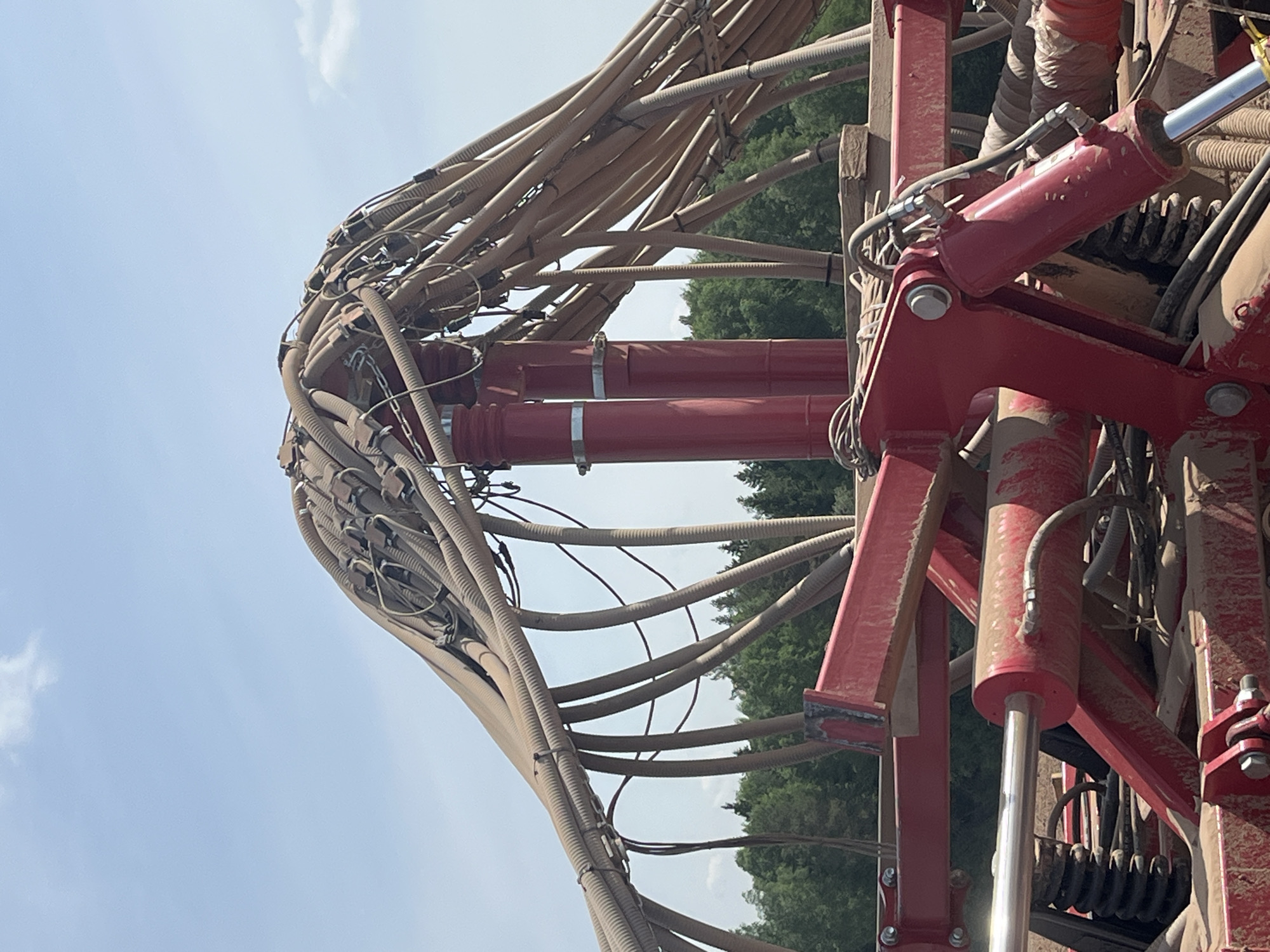The image depicts a complex arrangement of hoses and hydraulic cylinders on a piece of heavy machinery. The hoses are light brown and corrugated, bundled together with straps. Two red hydraulic cylinders are prominently visible, connected by metal brackets and hardware. The machinery appears to be in a rural setting, with trees visible in the background and a blue sky above. The equipment is dusty and shows signs of use. The location is noted as Unknown, Unknown.