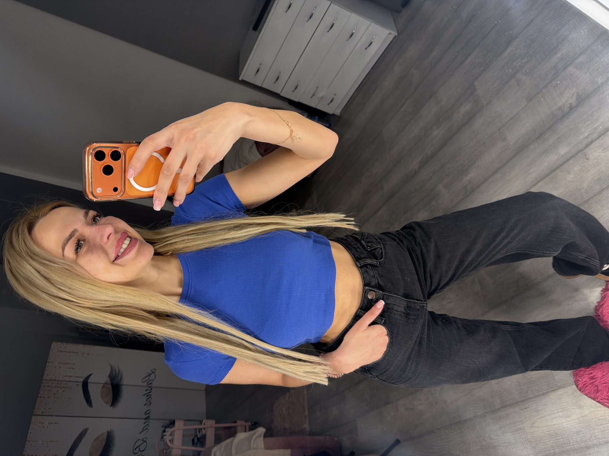 A blonde woman is taking a mirror selfie in a room. She is wearing a blue cropped t-shirt, black jeans, and fluffy pink slippers. She has her hand in her pocket and is holding a phone with an orange case. The room has a wooden floor and a dresser with multiple drawers. In the background, there is artwork of closed eyes and eyelashes on a wall. There is also a pink trolley with shelves visible on the left side of the image. The overall setting appears to be a bedroom or dressing area. The lighting is moderately bright, and the image is taken from a medium-low angle. The woman is smiling and looking at her reflection in the mirror.