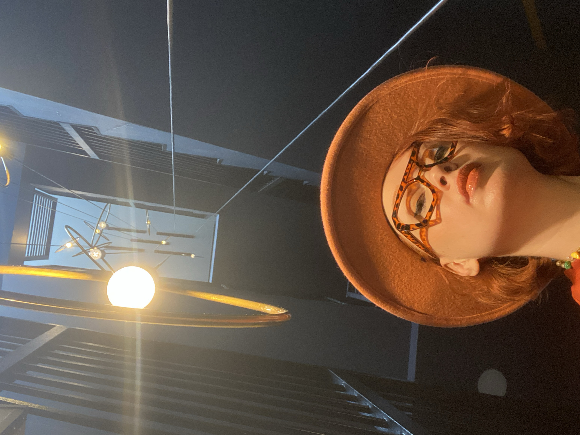 A low-angle shot depicts a person wearing an orange hat and orange-patterned glasses, gazing up towards a modern, multi-tiered building interior. The focal point is a bright, orb-shaped light fixture surrounded by a circular structure. The environment suggests a contemporary urban setting, possibly a public space or commercial building.