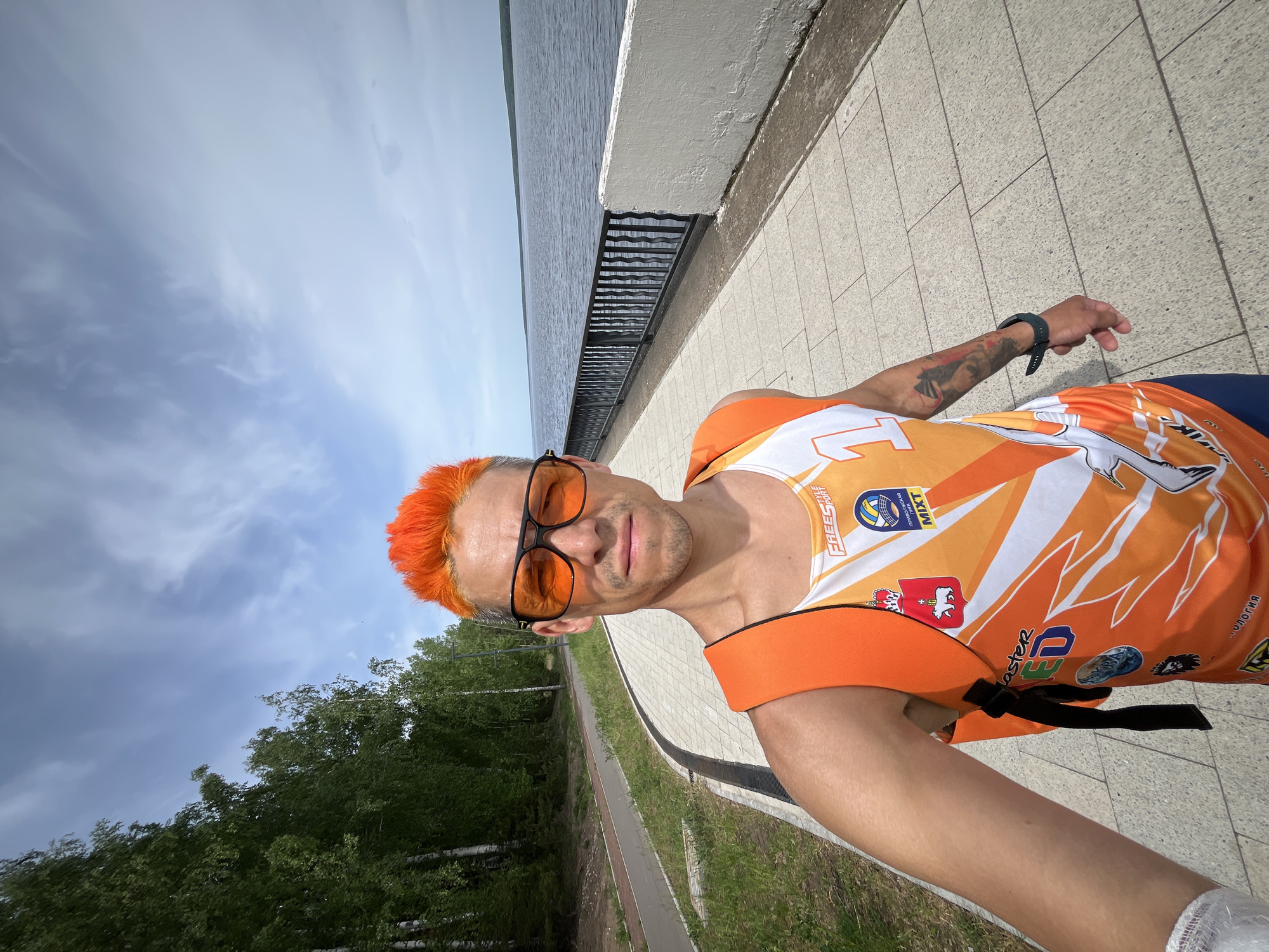 A man with orange hair and sunglasses is standing on a paved walkway. He is wearing an orange and white athletic tank top. A tattoo is visible on his left arm. The walkway is bordered by a low concrete wall and a black metal railing on one side, with water visible beyond the railing. On the other side of the walkway is a green grassy area with trees. The man is taking a selfie. The image was taken in the city of Chaikovsky, Russia.