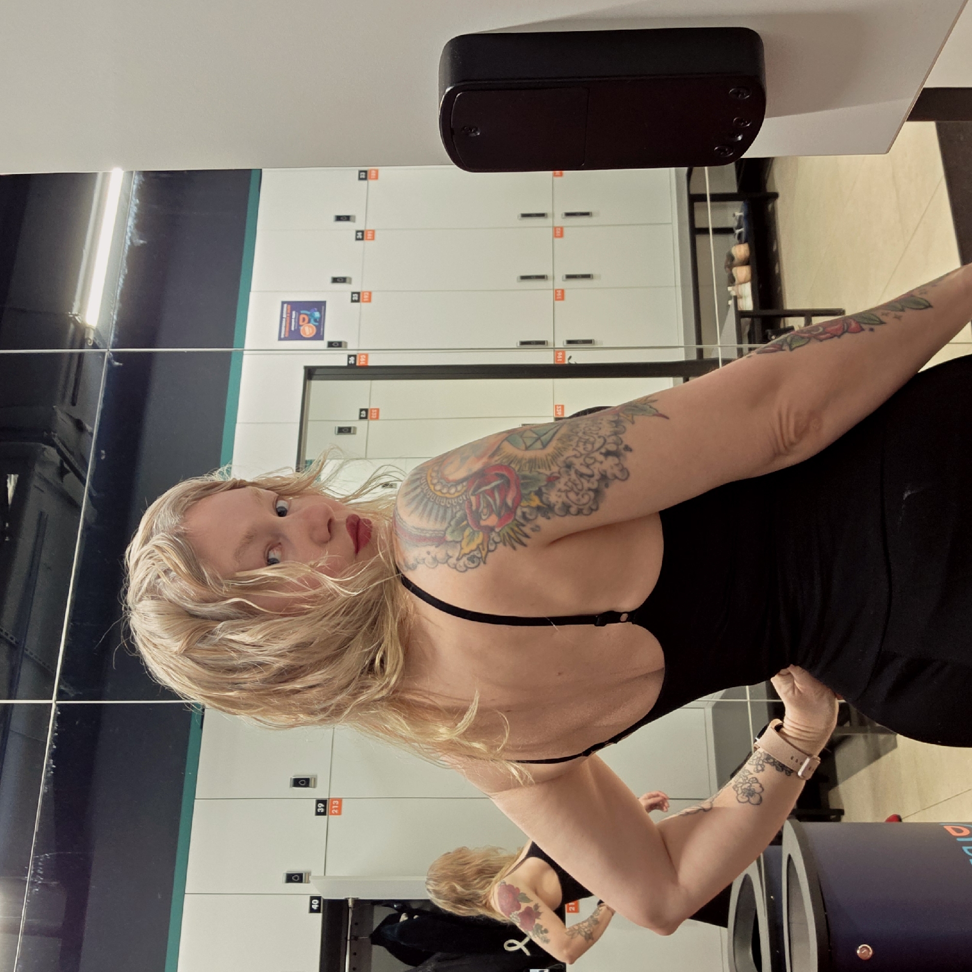 A woman with blonde hair, wearing a black dress and sporting several tattoos on her arms, poses for a selfie in front of a mirrored wall in a locker room. The locker room is equipped with rows of white lockers, each labeled with an orange number. A black rectangular object is mounted on the wall above the lockers. The location appears to be in the city of Saint Petersburg, Russia.