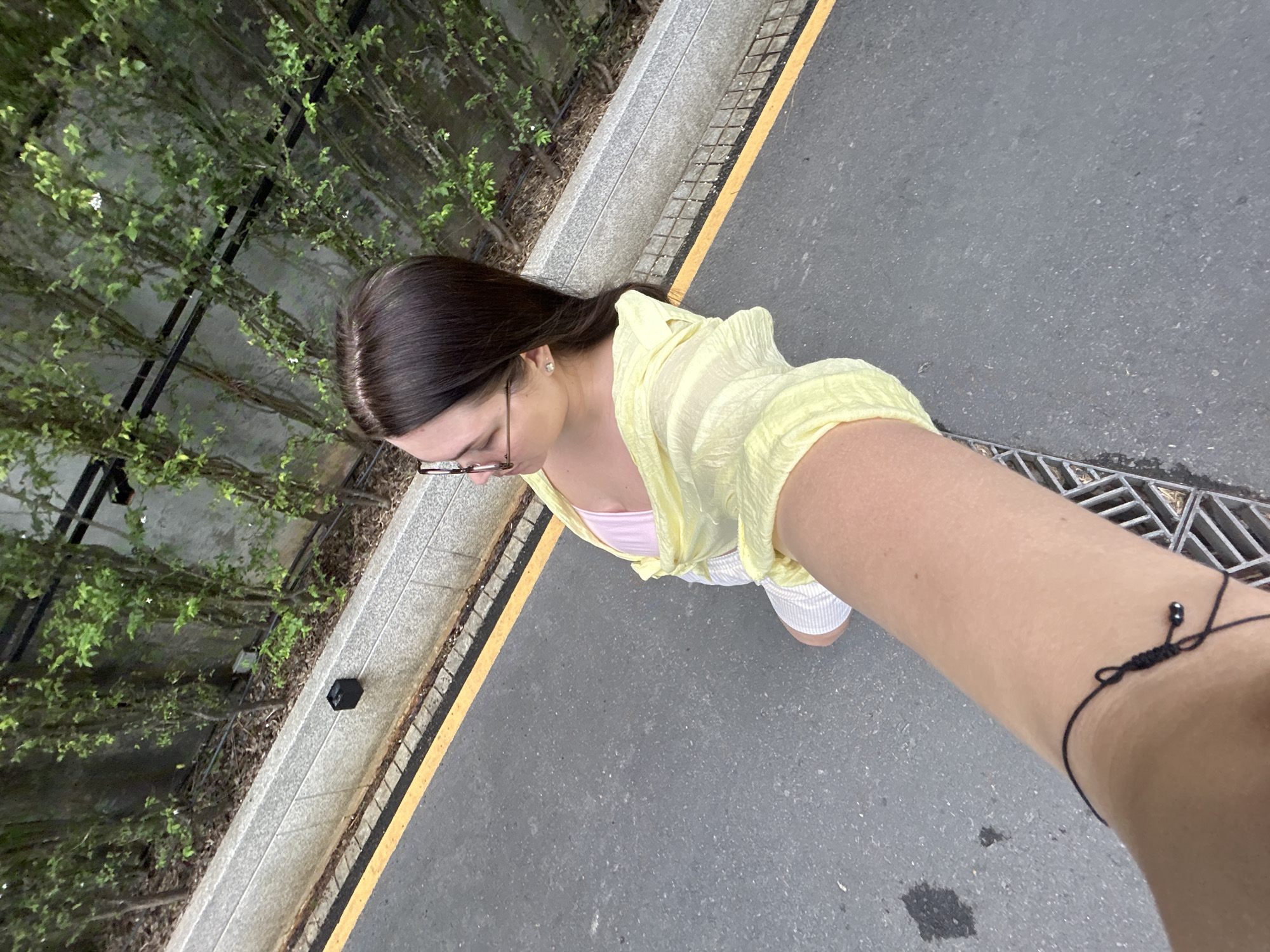 A high-angle shot depicts a person standing on a paved surface with a curb edged with a yellow line. Adjacent to the curb is a raised planter bed filled with dense green foliage. The person, seen from above, wears glasses, a pale yellow blouse, a pink top underneath, and white shorts. An arm extends into the frame, wearing a black cord bracelet. The setting appears to be an outdoor urban environment. The photo was taken in Rawai, Thailand.