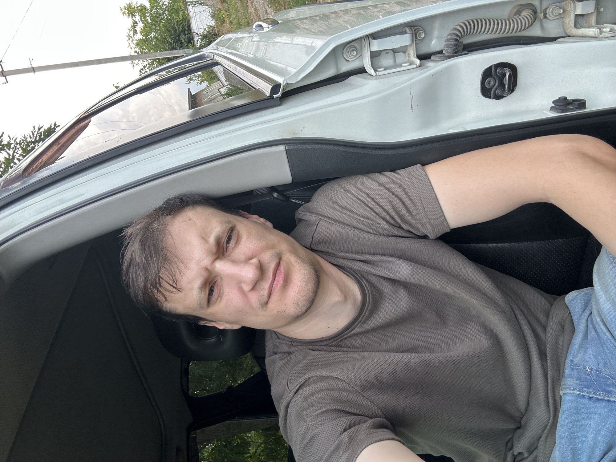 A man with short brown hair and a grey t-shirt is sitting in the driver's seat of a light-colored vehicle, with the driver's side door open. He is looking at the camera with a slightly furrowed brow and a neutral expression. The interior of the car is dark, with a black seat visible. The exterior shows a rural setting with trees and a utility pole. The image appears to be a selfie taken in Makiv, Ukraine, based on the provided context.
