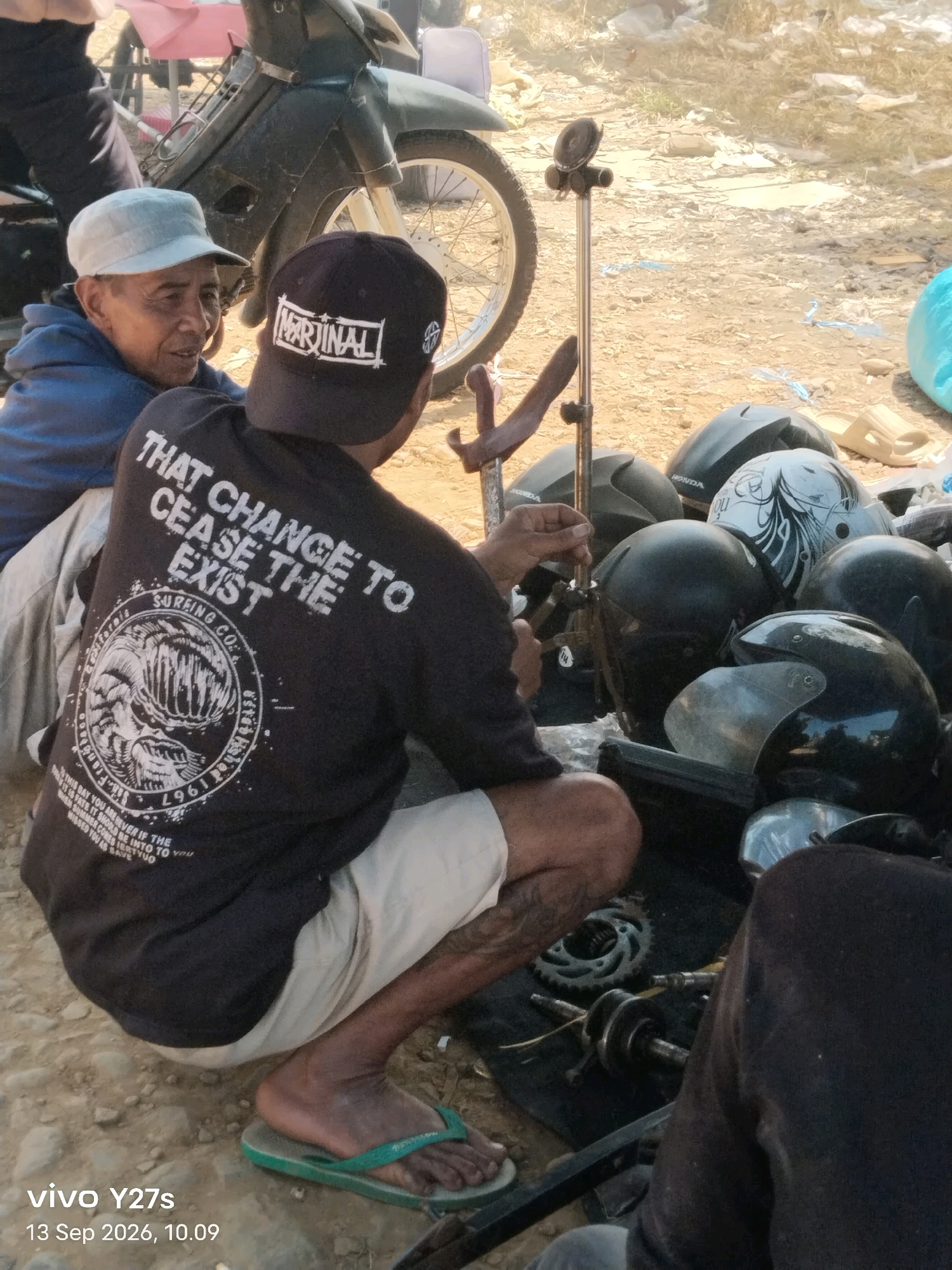 Two men are seated outdoors in Trimulyo, Indonesia. The man in the foreground, wearing a black t-shirt with white text and graphics, a black baseball cap, and light-colored shorts, is facing away from the camera and looking downwards. His right foot, adorned with a green flip-flop, rests on the ground. To his left, another man, wearing a blue jacket and a light-colored cap, is looking towards the man in the black t-shirt. They are surrounded by a collection of black motorcycle helmets, a motorcycle in the background, and various other unidentifiable objects on the dusty ground. The lighting suggests it is daytime.