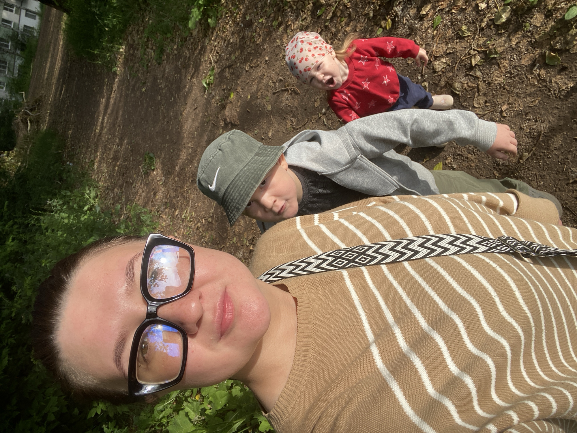 In Chaikovsky, Russia, a woman wearing large black-rimmed glasses and a brown and white striped sweater is taking a selfie outdoors. She is positioned on the left side of the frame. To her right, two children are visible. The child in the middle is wearing a gray bucket hat with a Nike logo and a gray hoodie over a dark shirt. The child on the far right is wearing a red long-sleeved shirt with white stars and a bandana with a cherry pattern. They are standing on a dirt path in a wooded area with green foliage on the left and a building visible in the background on the right. The ground is covered with dirt and fallen leaves, and sunlight is casting shadows. The woman has a bag with a black and white geometric strap slung across her chest. The children appear to be walking along the path behind the woman.