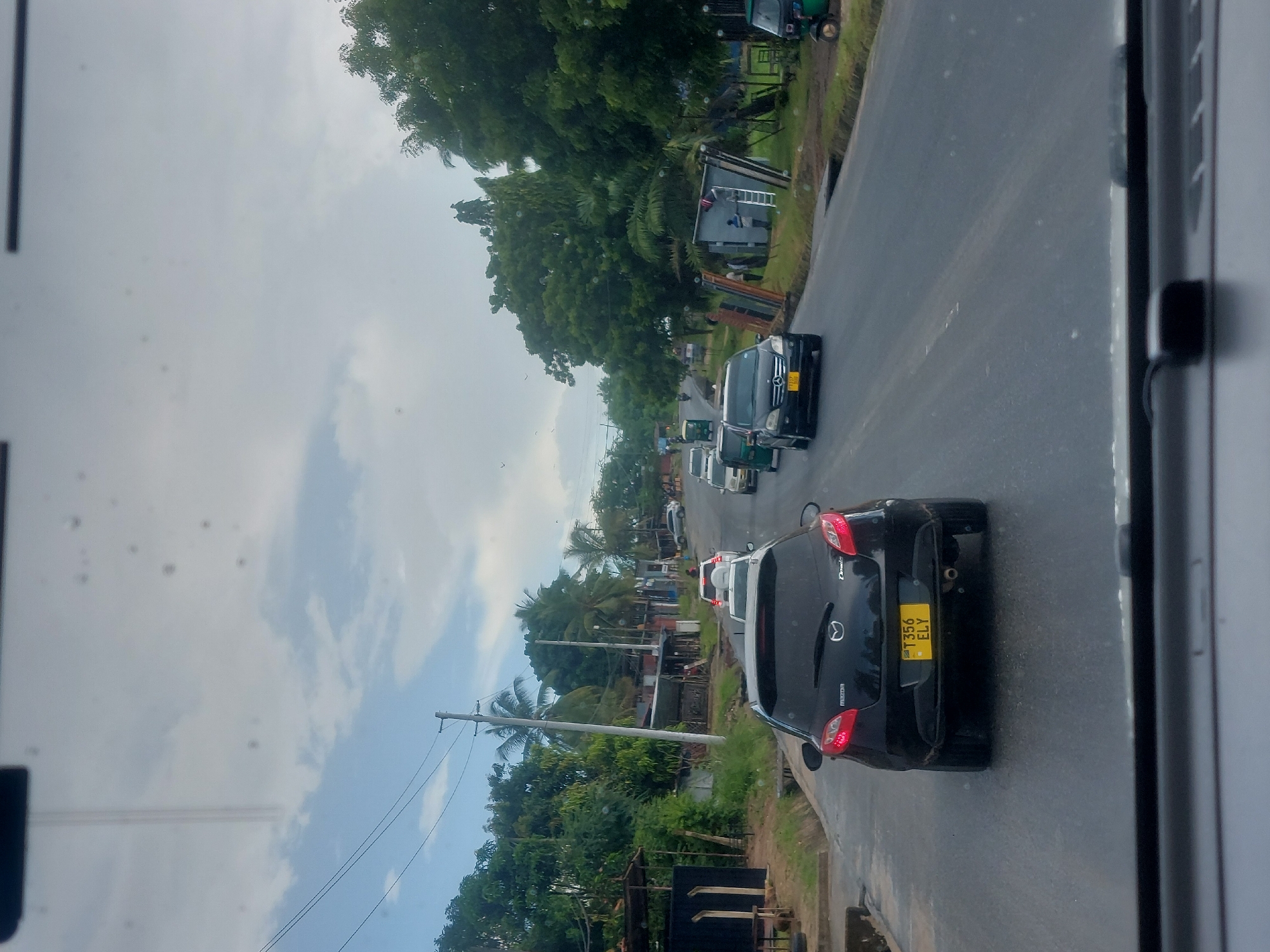The image shows a street in Dar es-Salaam, Tanzania. Several vehicles, including cars and a tuk-tuk, are on the road. A black Mazda is in the foreground, with the license plate "T356 ELY" visible. The environment includes trees, buildings, and utility poles. The sky is overcast. Some construction or renovation appears to be happening alongside the road.