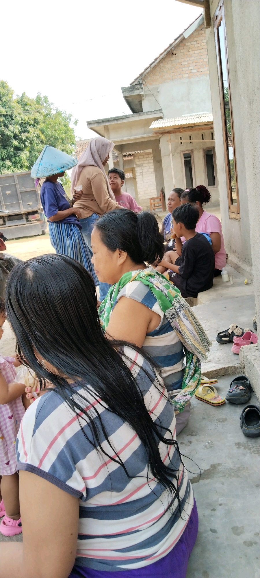 The image depicts a group of people gathered outdoors in front of a building under construction, likely in Tanjung Ratu Ilir, Indonesia. In the foreground, two women are visible from the back, one with long, dark hair wearing a striped shirt and purple pants. To their left, a young child in a pink dress with purple shoes is partially visible.
Behind them, several other individuals are present. A woman wearing a light brown hijab and a beige top is conversing with another woman in a blue shirt and a distinctive cone-shaped hat. Between them, a young boy in a pink shirt looks upwards. Further to the right, a group of women are seated on a ledge, including one in a pink t-shirt and another in a blue shirt. A young boy in a black t-shirt is also seated. Scattered on the ground are various pairs of children's shoes, including pink sandals, black clogs, and yellow flip-flops.
The environment suggests an informal gathering in a semi-rural or developing area. The building behind the people appears to be in the early stages of construction, with visible brickwork and exposed structural elements. Trees and foliage are present in the background, indicating an outdoor setting. The overall scene suggests a moment of social interaction amidst everyday life, possibly during a family visit or community event.