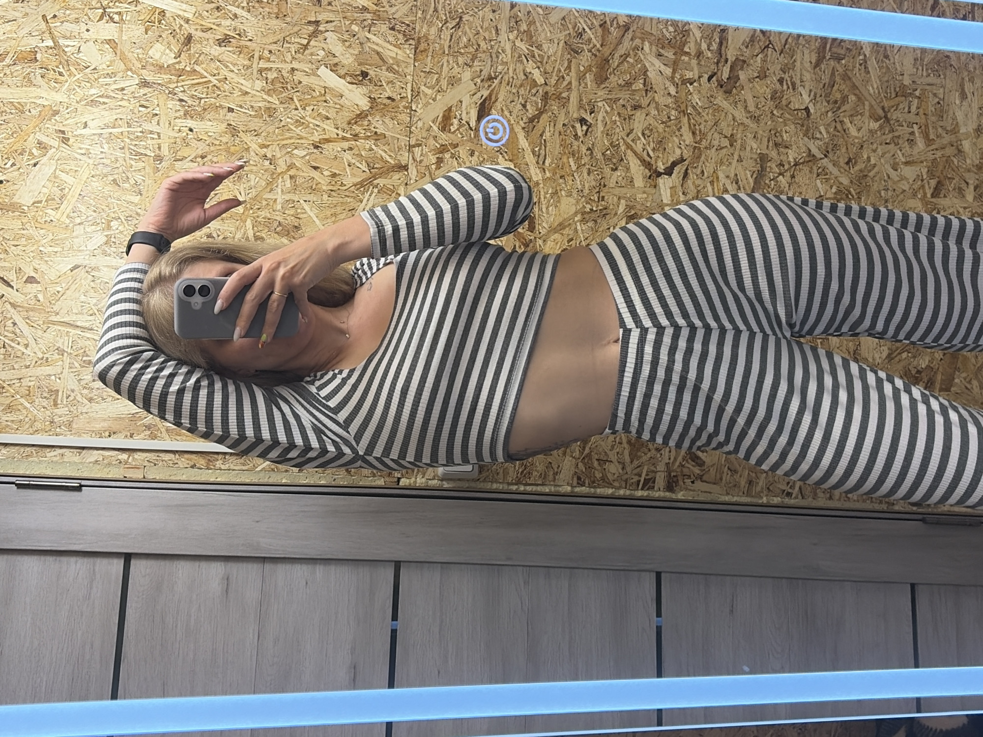 A woman is visible in a mirror. She is wearing a striped two-piece outfit. She is holding a smartphone up to take a picture of herself. The reflection shows a wall that appears to be made of wooden boards and another wall that is light gray and appears to be made of wood paneling. The location is not discernible from the image.