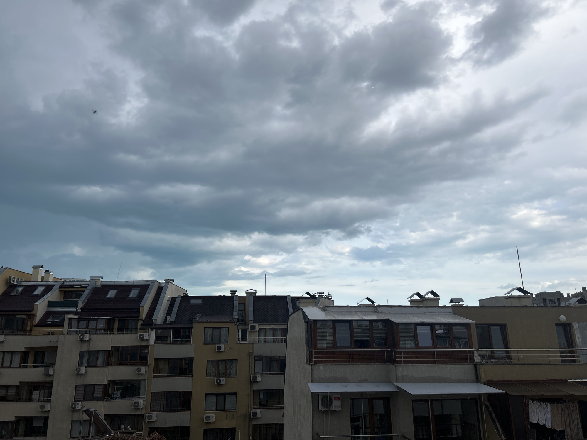 The image shows an urban landscape in Varna, Bulgaria. In the foreground are rooftops of multi-story residential buildings, predominantly constructed with light-colored facades and many windows and balconies. Air conditioning units are visible on the exterior walls of the buildings. The sky is overcast with heavy, grey clouds, suggesting inclement weather. A small, dark object, potentially a bird or an insect, is visible in the upper left portion of the sky. There are no people visible in the frame.