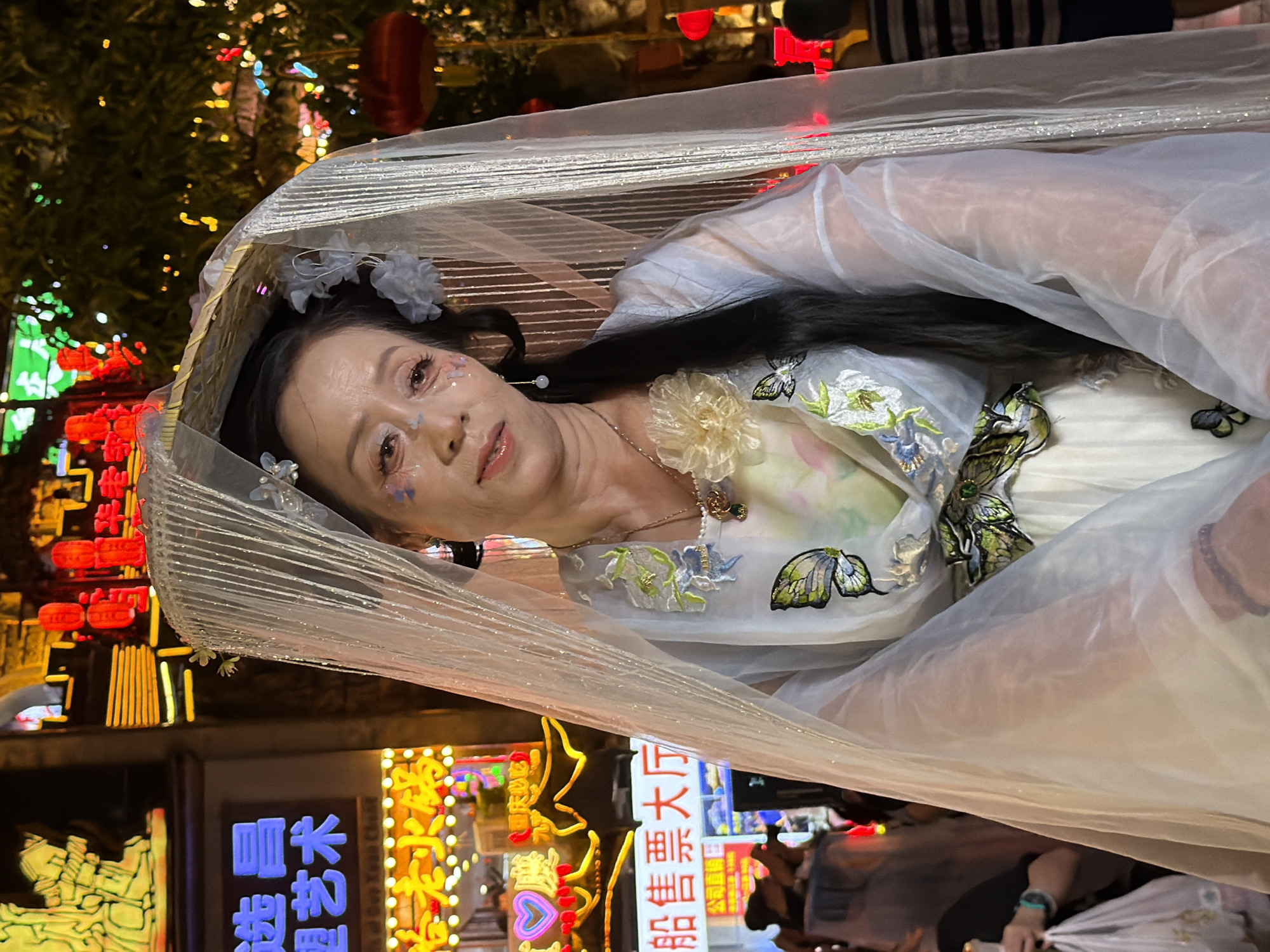 The media file shows a woman adorned in a traditional Chinese costume, possibly for a performance or festival. She is lying down, with a sheer, white, veil-like fabric draped over her. The woman has intricate makeup on her face, with glitter and light blue accents around her eyes, and small floral decorations in her hair. Her dress is white with embroidered floral and butterfly motifs.
The background suggests an outdoor setting at night, with numerous illuminated signs and lanterns. The vibrant colors of the neon signs and red lanterns indicate a lively urban environment, likely a commercial or entertainment district. Some of the visible text appears to be in Chinese characters, including what looks like "航售票大厅" (Háng shòu piào dà tīng), which could translate to "Ticket Hall". The overall atmosphere is festive and visually rich due to the abundant lighting. The woman's expression is serene, and the scene evokes a sense of theatricality or cultural celebration.