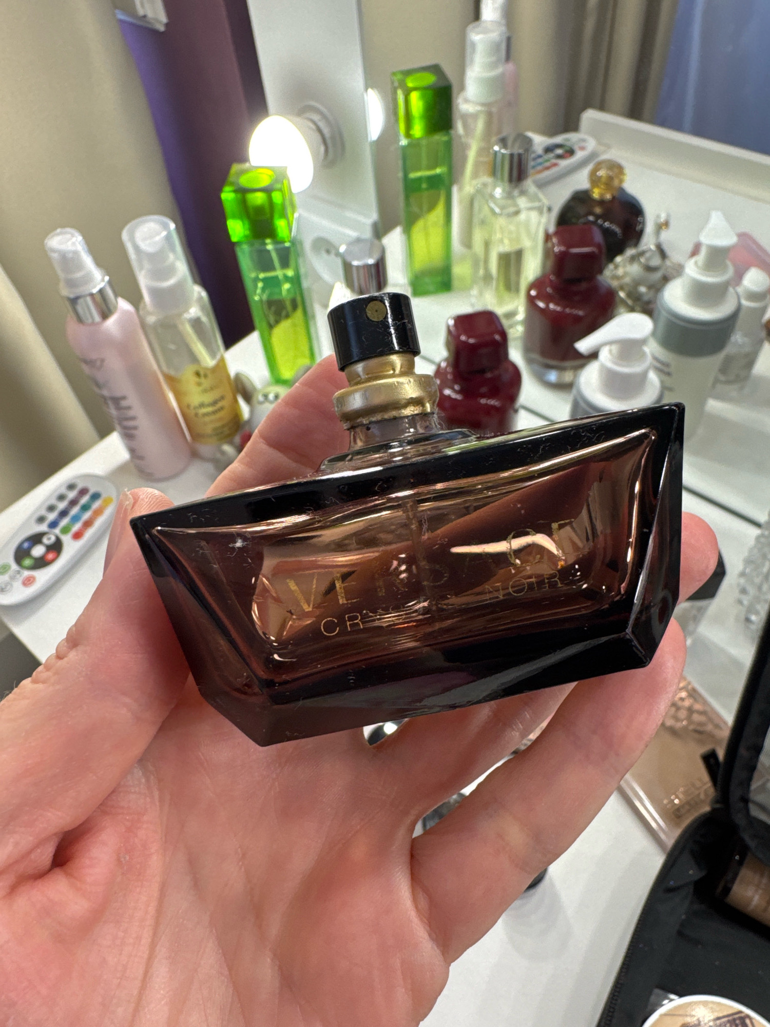 In a dimly lit room, a hand holds a dark brown glass perfume bottle with a gold and black cap. The bottle is partially filled with a light brown liquid and has "VE" and "CR NOIR" etched onto its surface. The background is cluttered with various cosmetic items, including spray bottles, glass bottles, and a remote control with colored buttons. The scene appears to be a personal vanity or dressing area. The image does not contain any information about the location in Sinyavino, Russia.