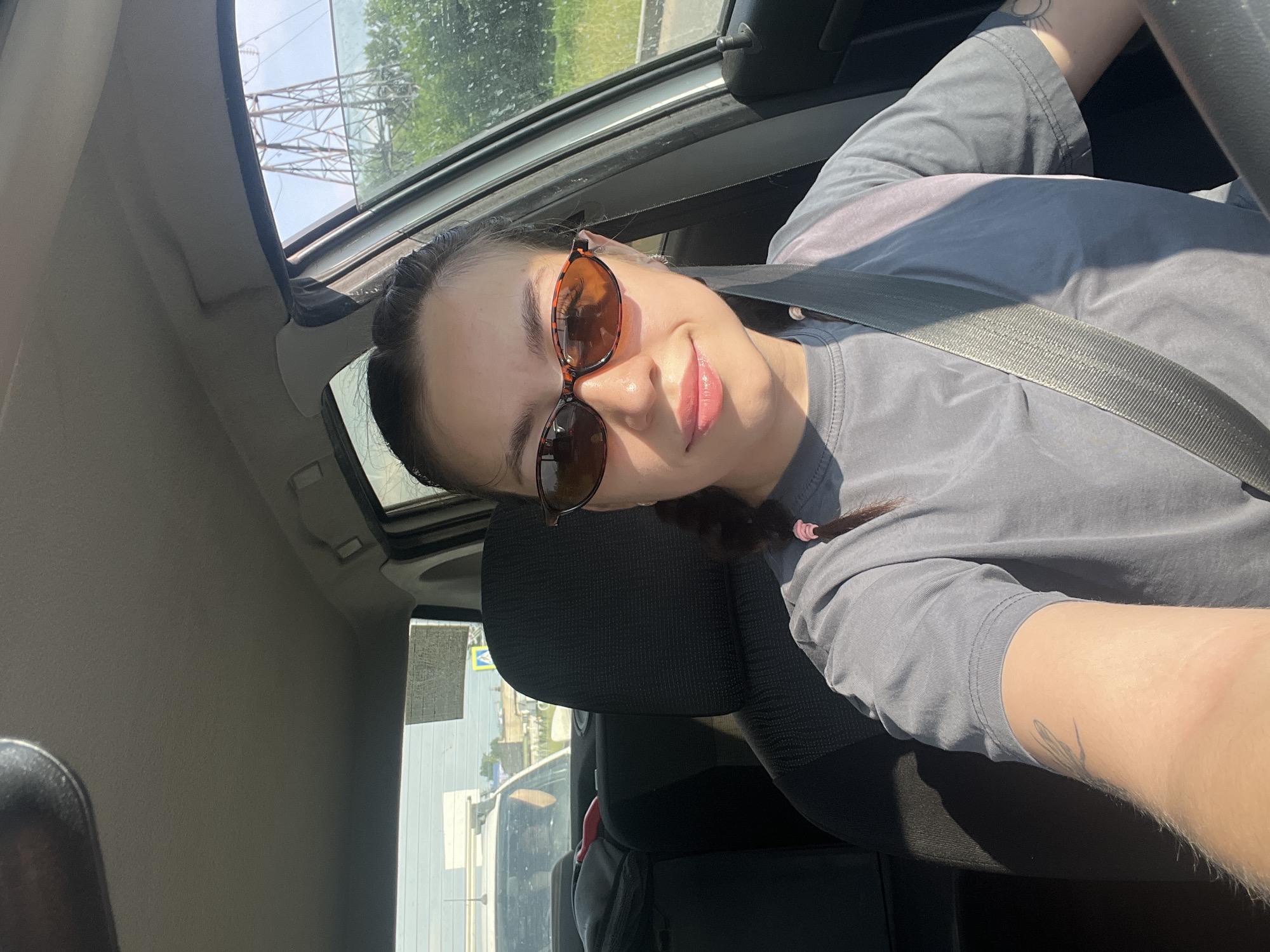 In a selfie taken from the passenger seat of a car, a young woman with dark hair pulled back in a braid wears sunglasses and a grey t-shirt. She is looking at the camera and smiling. The interior of the car is visible, including the seat, seatbelt, and dashboard. Outside the car window, trees and a power line tower are visible. The scene appears to be a sunny day in Chaikovsky, Russia, judging by the power line tower which is characteristic of Russian infrastructure.
