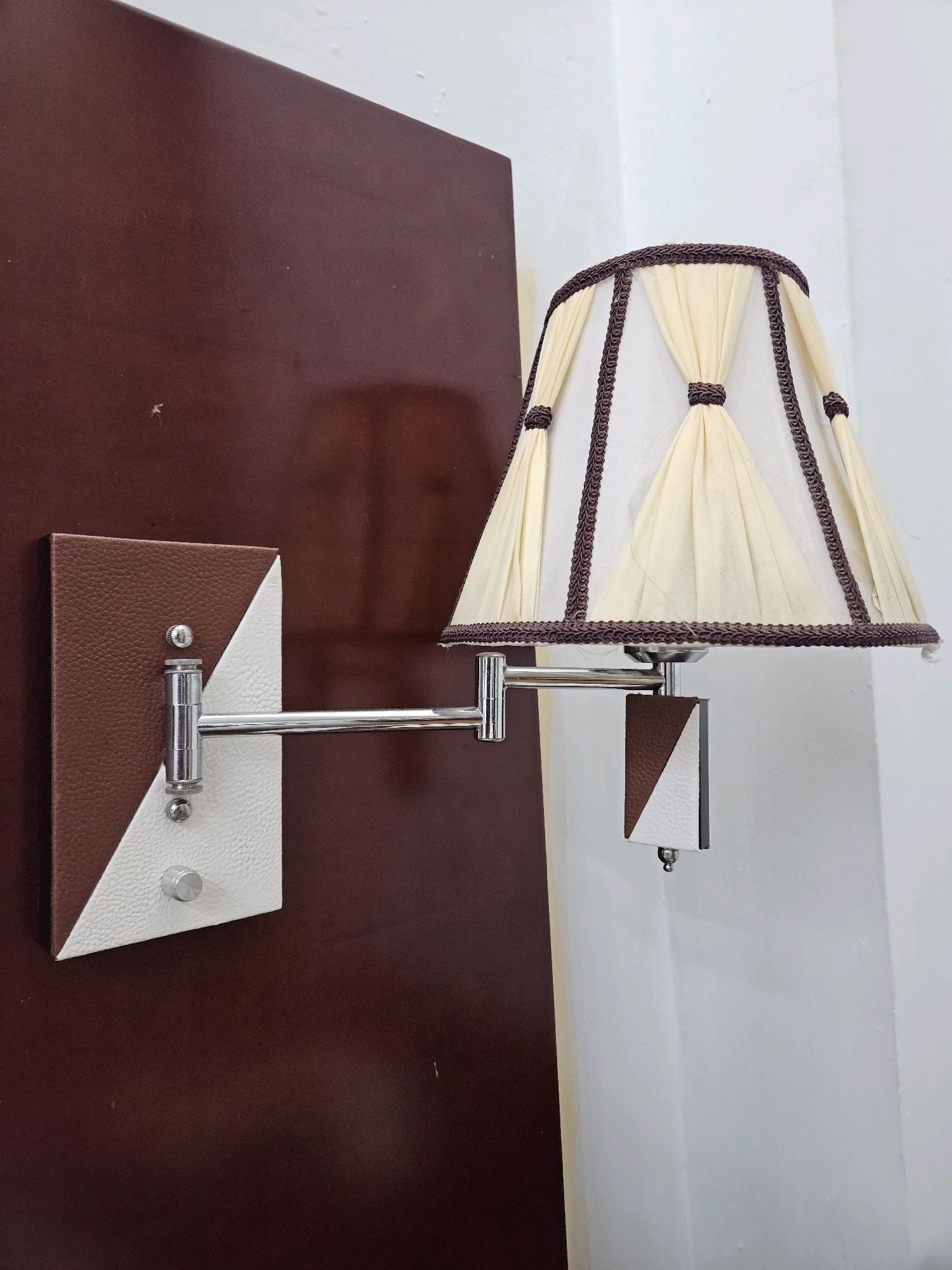 The image shows a close-up of a wall-mounted lamp. The lamp has a cream-colored fabric shade with decorative brown trim and tassels. The shade is supported by an adjustable silver-colored metal arm. The base of the lamp is attached to a dark red wall. The base itself is a square plate with two triangular sections: one brown and one white. There are two silver knobs on the base plate. To the right of the lamp, a white wall is visible. The scene is indoors, likely in a residential or hotel setting. There are no people or animals in the image, and no visible actions are taking place. The lighting appears to be natural or from an overhead source, creating subtle shadows. The image is taken from a slightly low angle, looking up towards the lamp. No specific location in Addis Ababa, Ethiopia can be identified from the visual information provided.