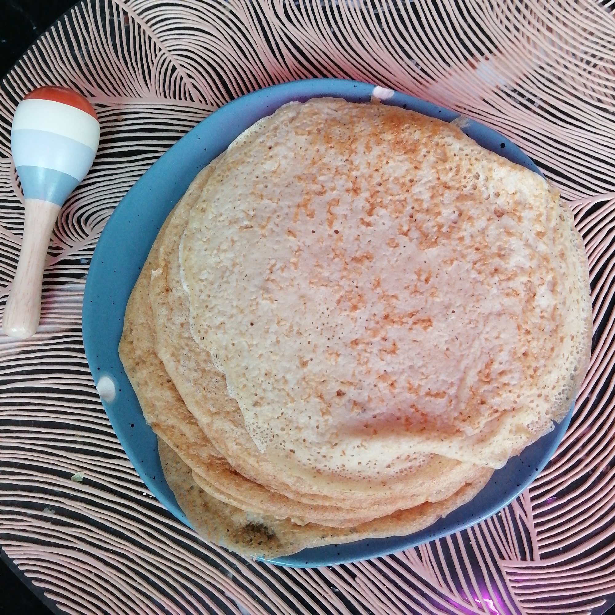 A top-down, close-up shot shows a stack of crêpes served on a blue plate against a black and pink abstract patterned background. To the left of the plate, there is a toy rattle with red, white, and blue bands. The crêpes are light brown with a slightly textured surface, stacked unevenly on the plate. The plate has a speckled, matte finish. The background pattern consists of wavy, parallel pink lines on a black surface. The lighting is diffused and even, with no strong shadows.