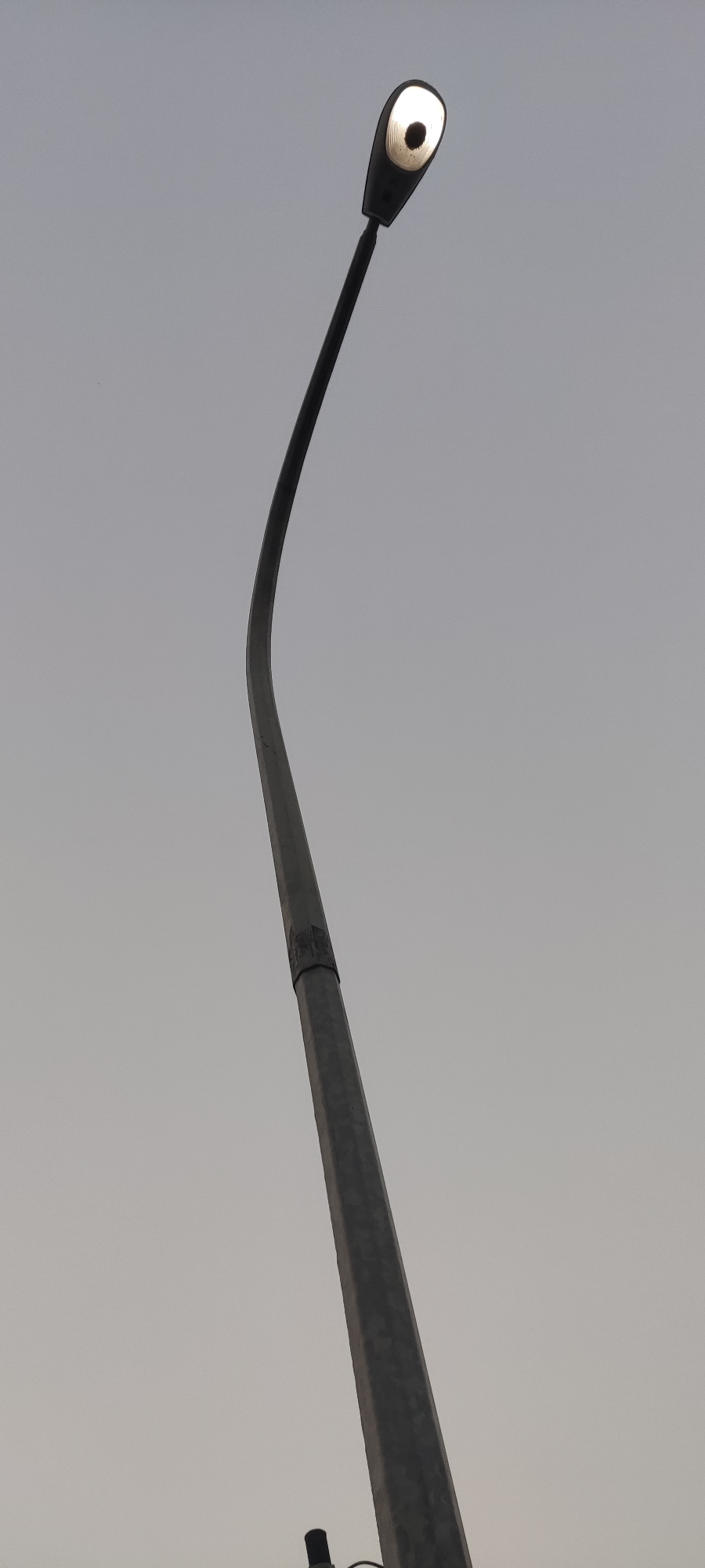 An outdoor, low-angle, vertical shot shows a street lamp against a pale gray sky. The lamp, illuminated with a bright white light, has a dark, oval-shaped lens and is mounted on a slender, black, curved arm. The arm extends from a thick, gray, octagonal pole that is angled towards the lower right of the frame. A dark band, possibly a joint or fixture, is visible on the pole. The bottom of the pole is cropped out of the image, but a small, dark cylindrical object is partially visible at the very bottom. The sky is overcast and appears to be twilight or a heavily clouded day. The scene is devoid of any other discernible objects or features, emphasizing the starkness of the lamp and pole against the uniform sky. The location of Pakusamben, Indonesia, is noted.