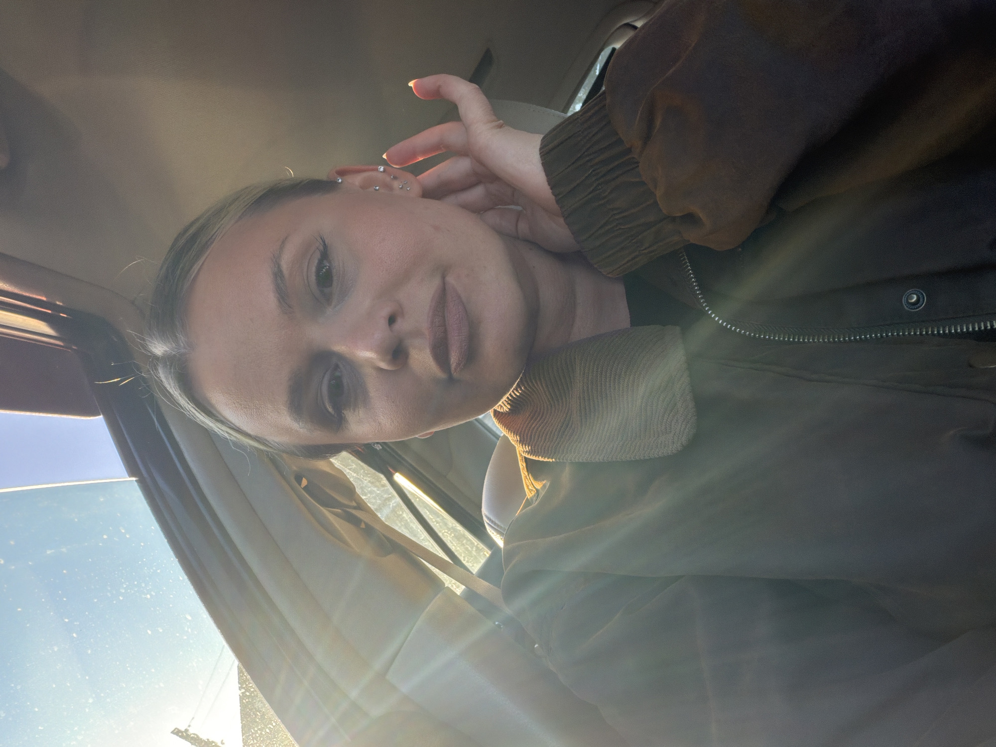 A woman with blonde hair is positioned inside a vehicle, likely a car, given the view of the windshield and the interior elements. She is wearing a brown jacket. Her hand is raised near her ear, which has multiple piercings. The lighting suggests it is daytime, with sunlight streaming into the vehicle through the windshield. The location is not determinable as there is no identifiable information about the city of Sarapul, Russia.