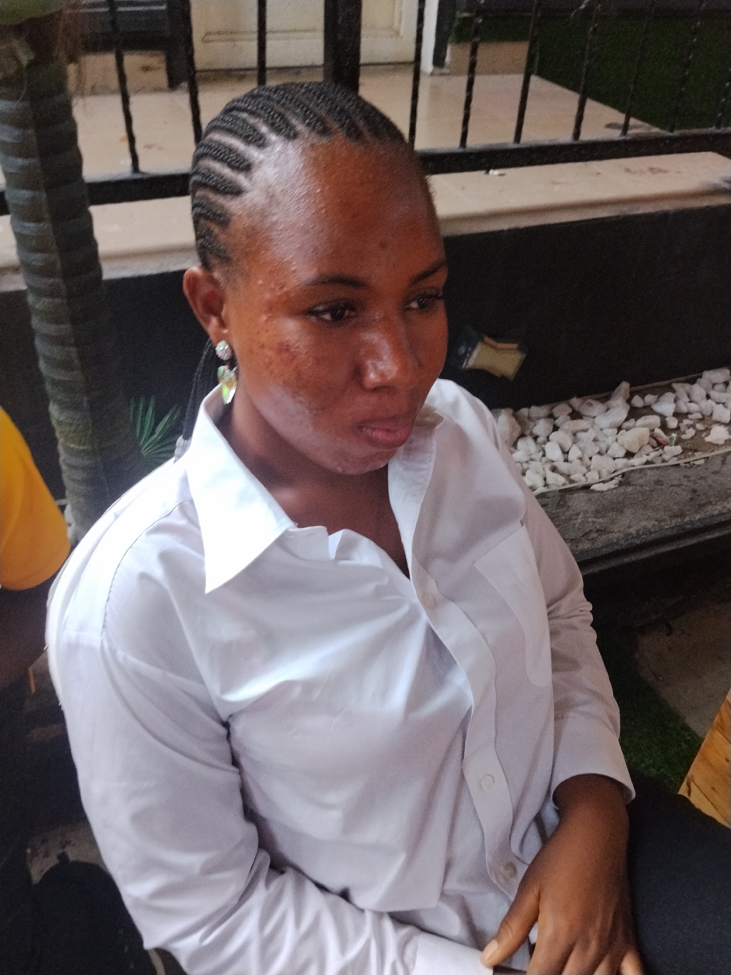 A medium shot, eye-level, full-body shot captures a young Black woman in a light-colored button-down shirt and dark pants. She is seated, looking to the right of the frame with her head tilted down. Her cornrowed hair is neatly styled, and she wears dangling earrings. The background includes a black metal railing, a palm tree, white pebbles on a ledge, and some green astroturf. The scene appears to be an outdoor seating area, potentially a restaurant or lounge, in Howayi, Nigeria. The lighting is natural, suggesting an overcast day or shaded area.