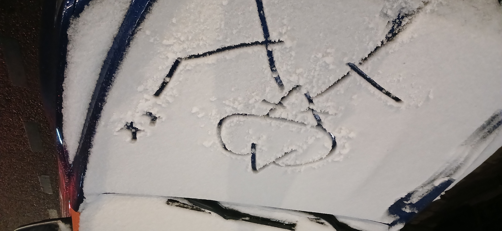 A blue car hood is covered in snow, with childish drawings etched into the snow. The drawings include a stick figure, a spade symbol, and a plus sign. The car is parked outdoors, possibly in a residential area. The location is not discernible from the image.