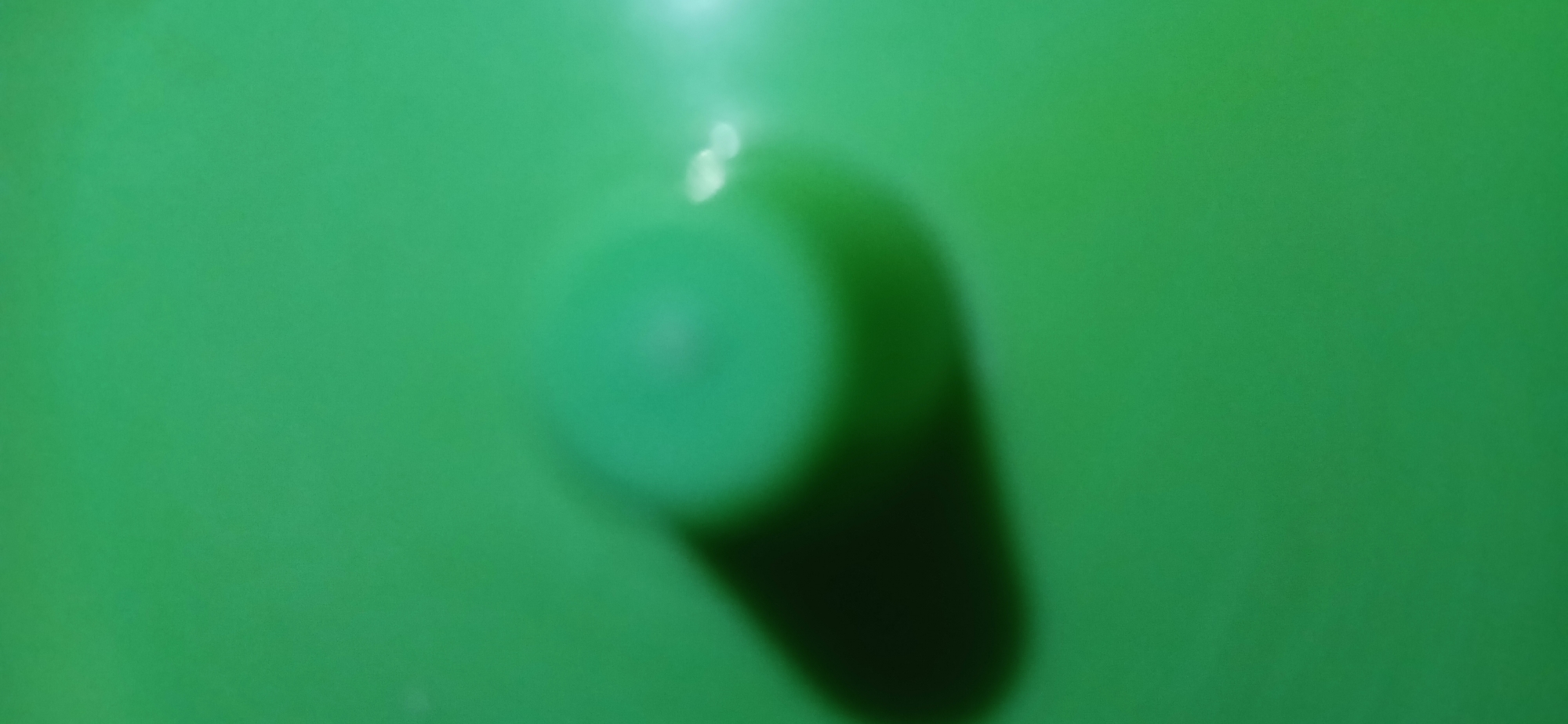 A blurred, close-up, overhead shot captures a green, textured surface. A circular object, also green but with a lighter center, is visible, casting a dark, elongated shadow to its left. The object appears to be sitting on the green surface, which could be a tabletop or a piece of fabric. There are also some bright white light reflections on the right side of the image. The image does not contain any recognizable landmarks or context to determine its location in Sekarputih, Indonesia.
