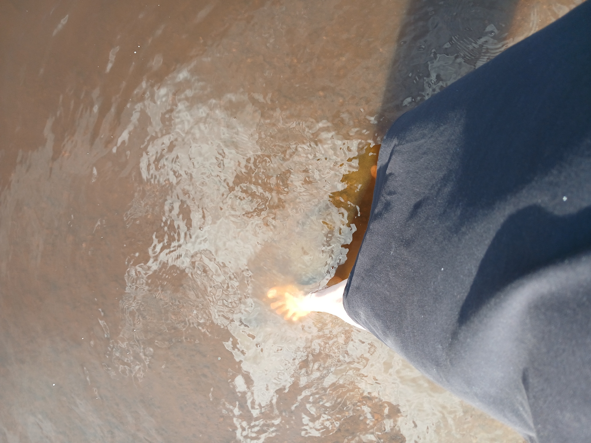 A bare foot, illuminated by sunlight, is submerged in shallow, murky water. The foot, appearing pale with visible toes, is positioned towards the lower left of the frame. A dark-colored garment, possibly a skirt or pants leg, covers the lower portion of the image, obscuring the rest of the person's body. The water's surface is disturbed, showing ripples and reflections, indicating movement. The bottom beneath the water is a mix of sand and small stones. The overall scene suggests an outdoor environment, potentially a riverbank or shallow body of water. The image does not provide any specific clues to its location within Yekaterinburg, Russia.