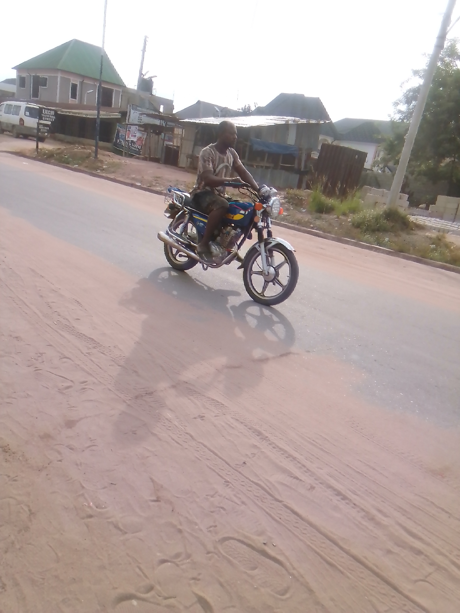 The image depicts a man riding a motorcycle on a paved road. The man is dark-skinned and dressed in a patterned shirt and dark shorts. The motorcycle is blue and yellow with chrome accents. The location is Owerri, Nigeria, based on the street signage in the background and the general environment, which is a semi-urban area with a mix of modern and traditional buildings. The road surface shows tire tracks and a sandy texture. Other buildings and a van are visible in the background, indicating a populated area. There is a utility pole on the right and a building with a green roof on the left.
