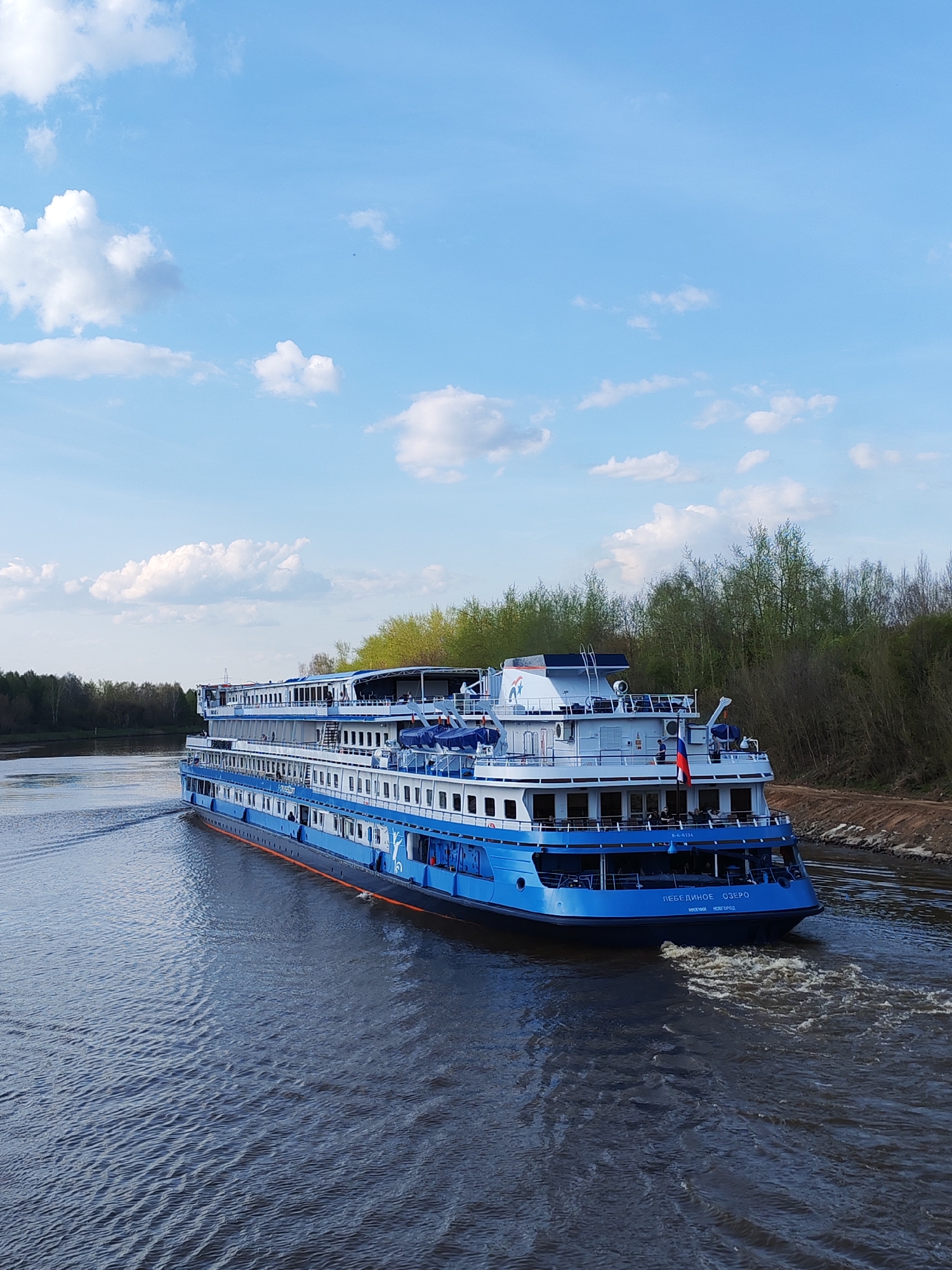 A large blue and white river cruise ship is traveling on a body of water in Varvarino, Russia. The ship, labeled "Лебединое Озеро" in Russian and the serial number "R-4-6134", creates a foamy wake. The river is bordered on both sides by green trees and shrubs. The sky is bright blue with scattered white clouds.
