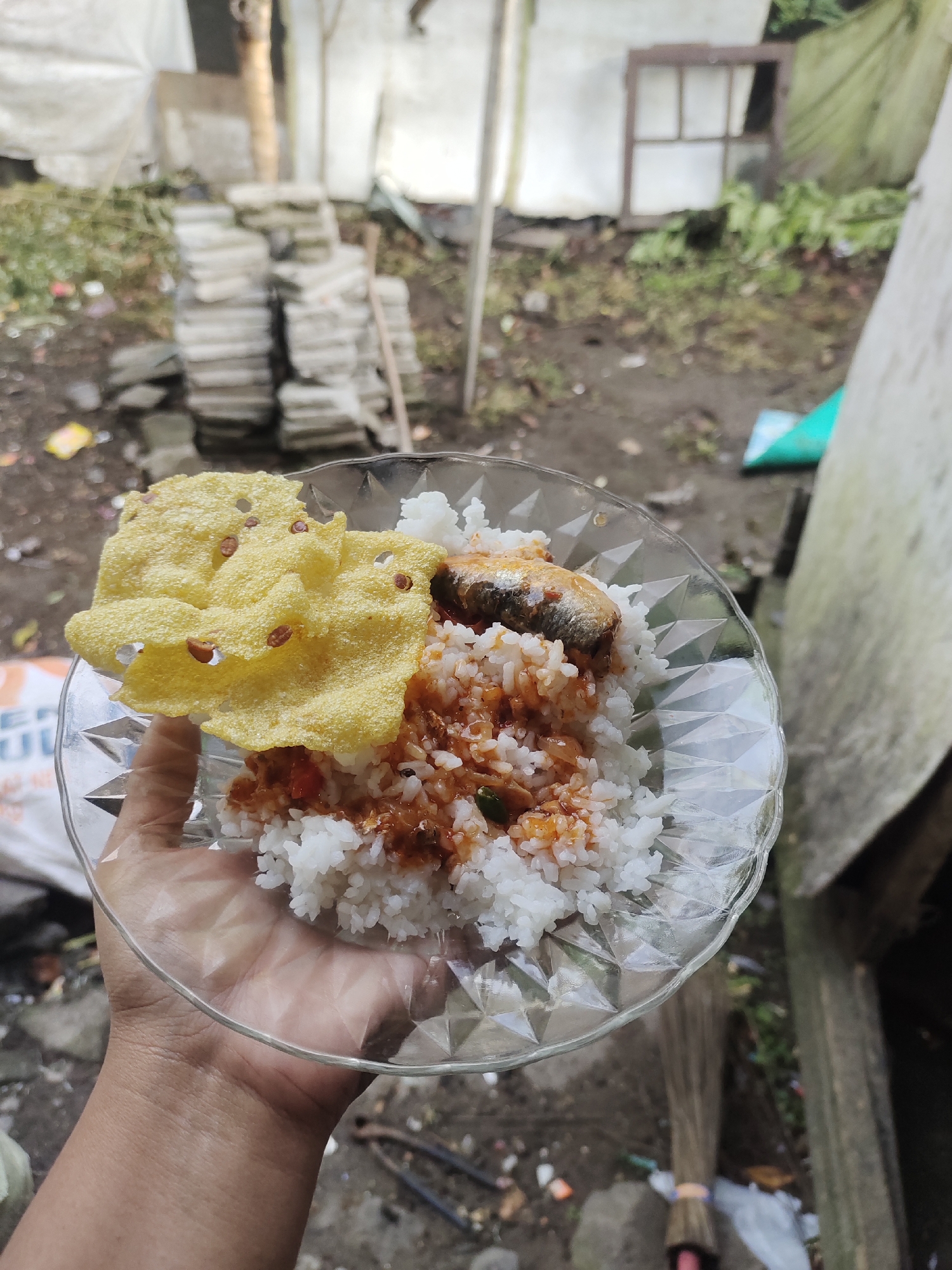 A person's hand holds a clear, patterned glass plate filled with white rice topped with a fish in sauce and a yellow cracker. The background suggests an outdoor setting with dirt, scattered debris, stacks of grey rectangular objects, some greenery, and a portion of a wooden structure. The scene appears to be in an outdoor, possibly rural or semi-developed area, consistent with a location in Cintadamai, Indonesia.