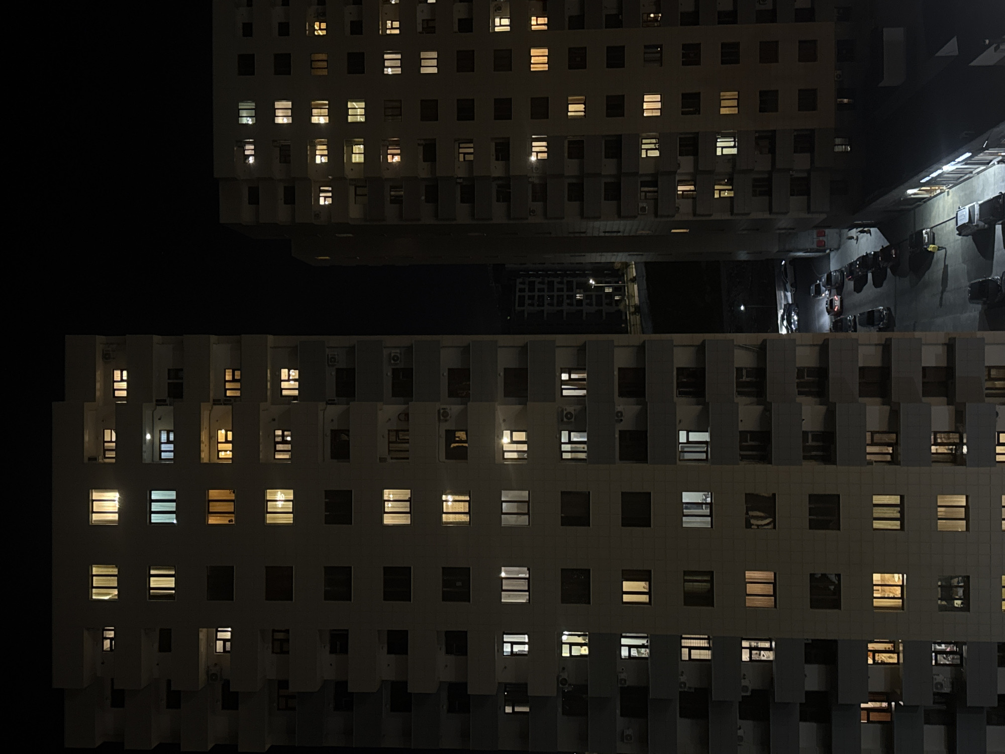 The original image presents a top-down, slightly oblique view of two modern buildings in Yerevan, Armenia, at night. The structures, likely residential or commercial, feature a repetitive grid of square windows across their light-colored facades. Many windows glow with warm, yellow-orange light from activated interior lights, while some emit cooler white or blue light, and others remain dark. Both buildings display a distinctive modular architecture with numerous rectangular projections, possibly balconies or architectural elements, many of which house visible air conditioning units.
The upper building is seen from a greater distance and height, while the lower building fills a significant portion of the foreground. A deep, dark void lies between them, suggesting a large open space or street. Below the upper building, a street or parking area is dimly illuminated by a few distant lights, revealing several cars parked along its edge. No people are discernible in the scene. The image portrays a tranquil urban night, highlighted by the geometric patterns of the architecture and the contrasting glow of scattered lights against the deep black of the night sky. No text is visible.