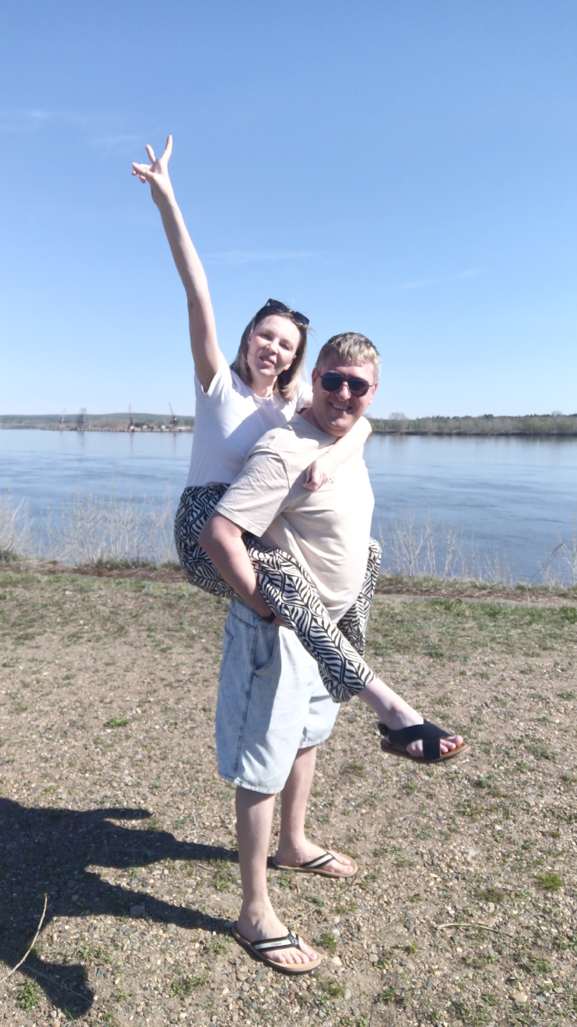 Two people are in the foreground. The location is along a riverbank.
One person is giving another a piggyback ride. The person giving the piggyback ride is a fair-skinned male with short blond hair. He is wearing sunglasses, a tan t-shirt, and light denim shorts. He is wearing brown flip-flops.
The person being carried is a fair-skinned female with blond hair, partially obscured by sunglasses worn on her head. She wears a white t-shirt and black-and-white patterned pants. She is wearing a single black sandal. Her left arm is raised, displaying the peace sign.
The background contains the river, across which land and vegetation are visible. Utility poles are visible across the river. The sky is clear blue. The ground is gravel.