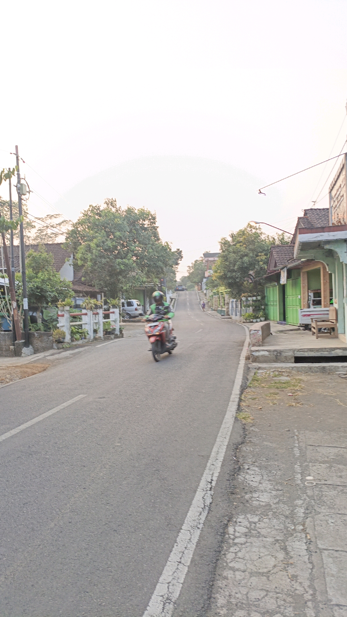 A person wearing a green helmet and jacket is riding a red scooter on a paved road in Salatiga, Indonesia. The road is lined with houses and trees on both sides. The sky is bright and overcast. There are some utility poles with wires on the left side of the road. The scooter rider is in motion, indicated by the blur. To the right of the road, there is a sidewalk with some vegetation and a drainage grate. Further down the road, there are more houses and trees. The scene appears to be a residential street in a rural or suburban area.