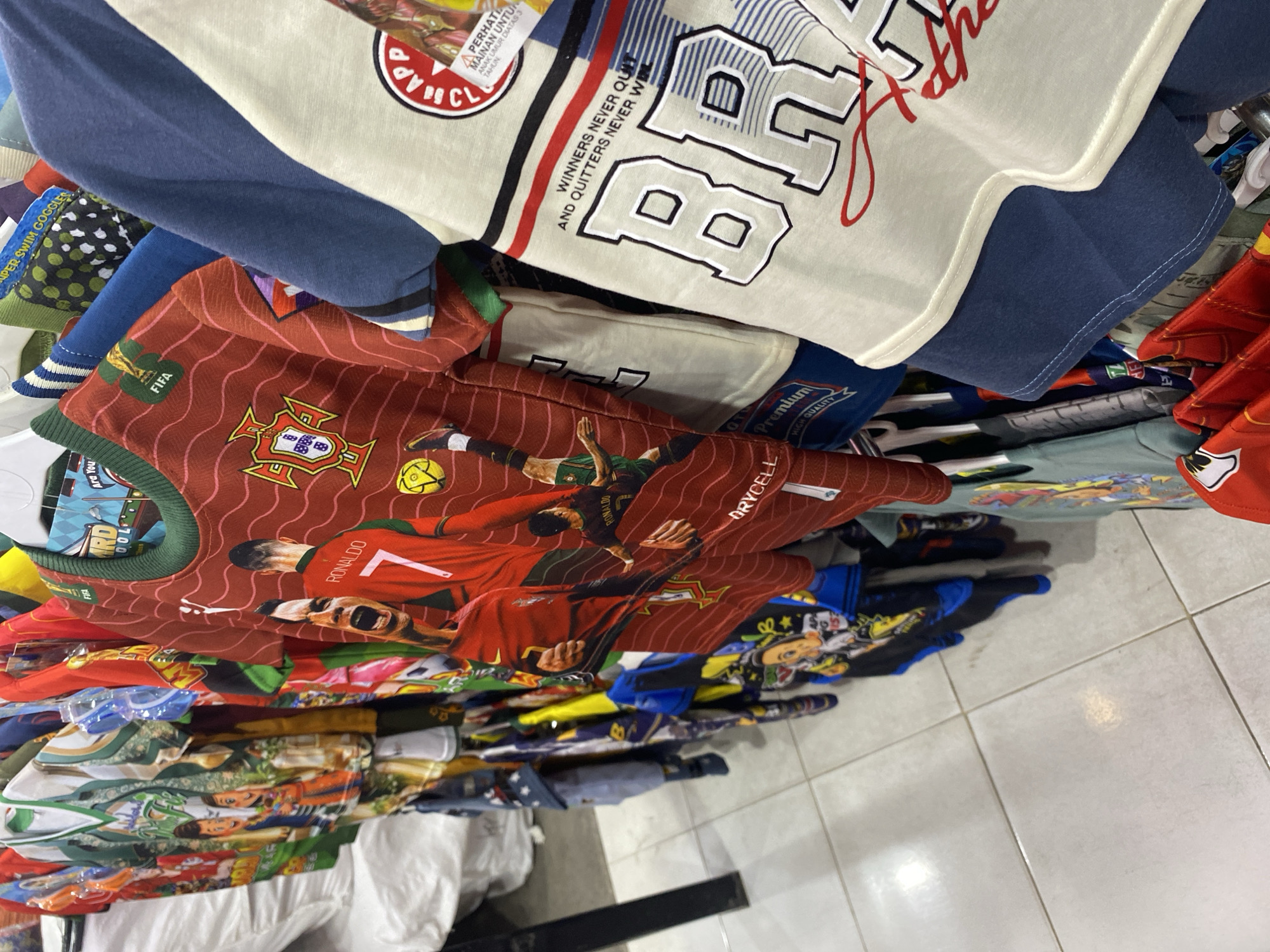 A clothing store in Banda Aceh, Indonesia displays a rack of t-shirts with vibrant designs. The t-shirts feature images of soccer players like Cristiano Ronaldo, along with team crests and motivational slogans such as "Winners Never Quit And Quitters Never Win." Some shirts are red with wavy patterns, while others are off-white with bold black and blue lettering. The background shows other clothing items on hangers and a tiled floor. The store appears to be well-stocked, with a variety of colorful apparel available for purchase.