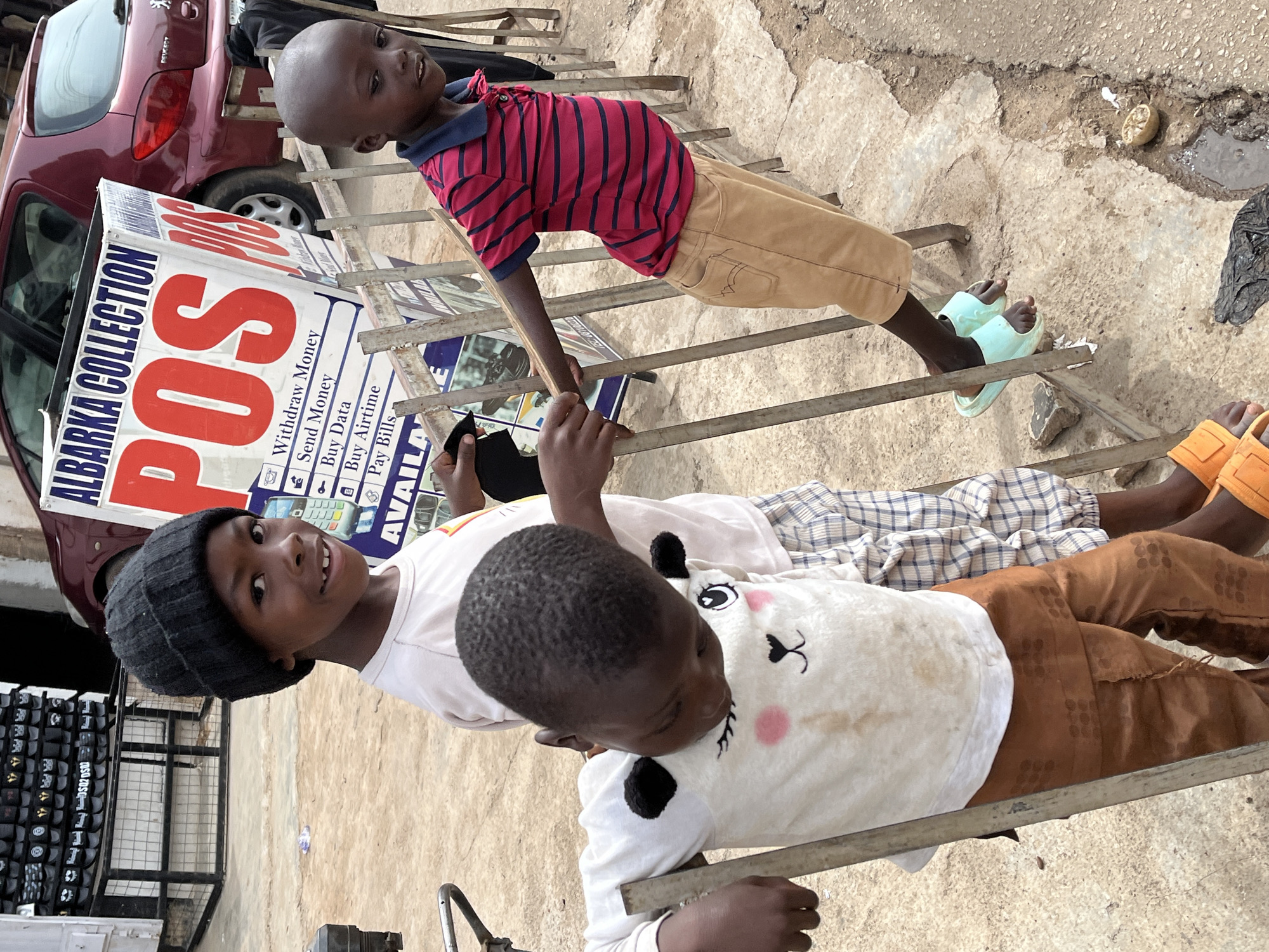 Three boys are seated on a ladder adjacent to an "ALBARKA COLLECTION POS" sign, near a red Peugeot vehicle. The boy at the top wears a red and blue striped shirt, khaki pants, and light green sandals. The middle boy has a black beanie, a white t-shirt, and plaid pants, smiling at the camera. The third boy is wearing a white t-shirt with a cartoon image and brown pants. The location appears to be in Jos, Nigeria, given the business sign.