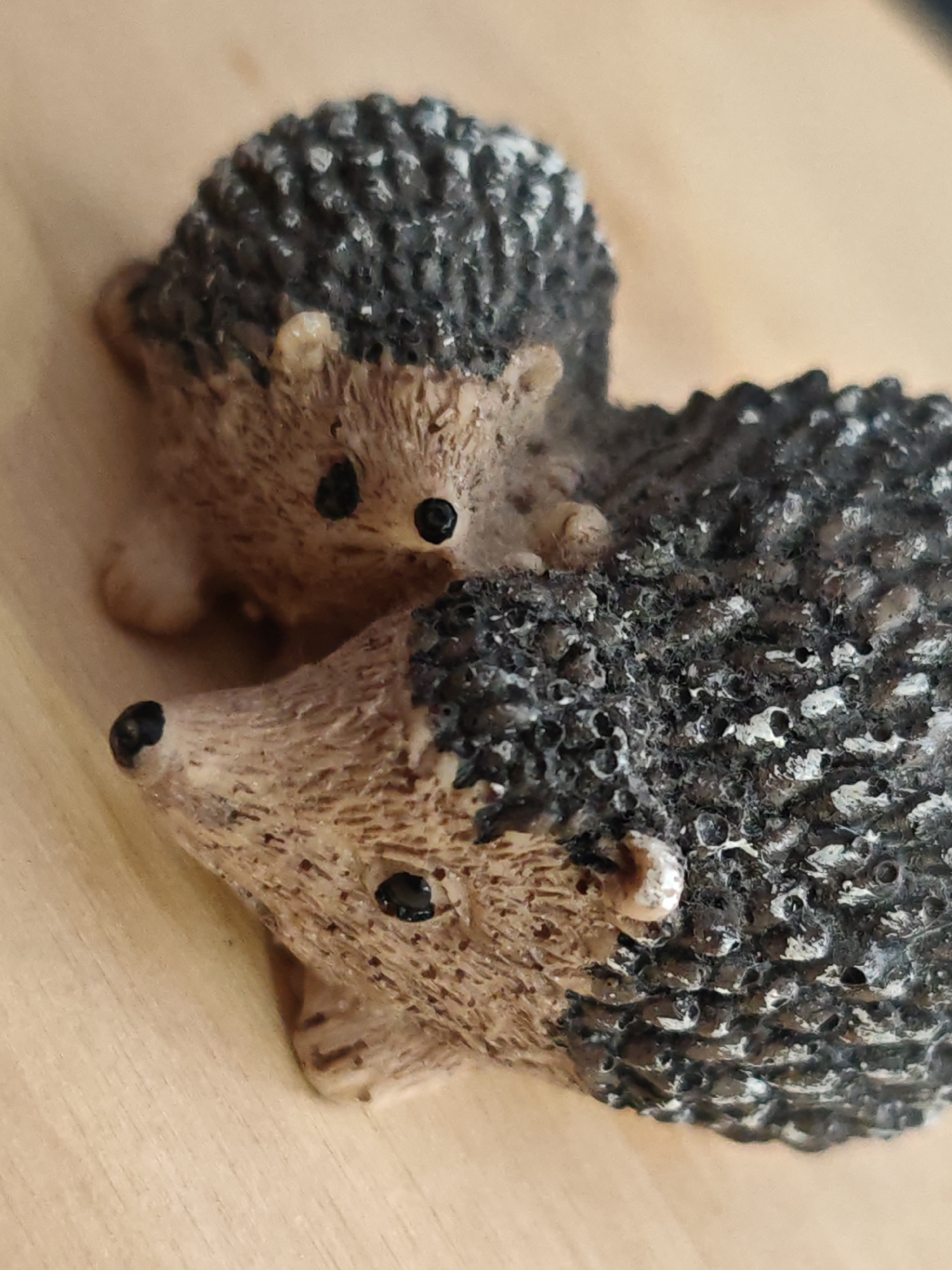 Two miniature hedgehog figurines are positioned on a light-colored, wooden surface. The larger figurine is in the foreground, facing left with its nose pointing towards the bottom left corner of the frame. Its back is textured to resemble quills, colored in shades of dark brown and black with lighter speckles. The face is a lighter brown, with a black nose and eye. Partially obscured behind the larger figurine is a smaller hedgehog, facing forward and slightly to the right. This figurine also has a textured, quill-like back in dark brown and black, and a lighter brown face with black eyes and nose. The wooden surface appears smooth with a visible grain. The lighting suggests it is an indoor setting.