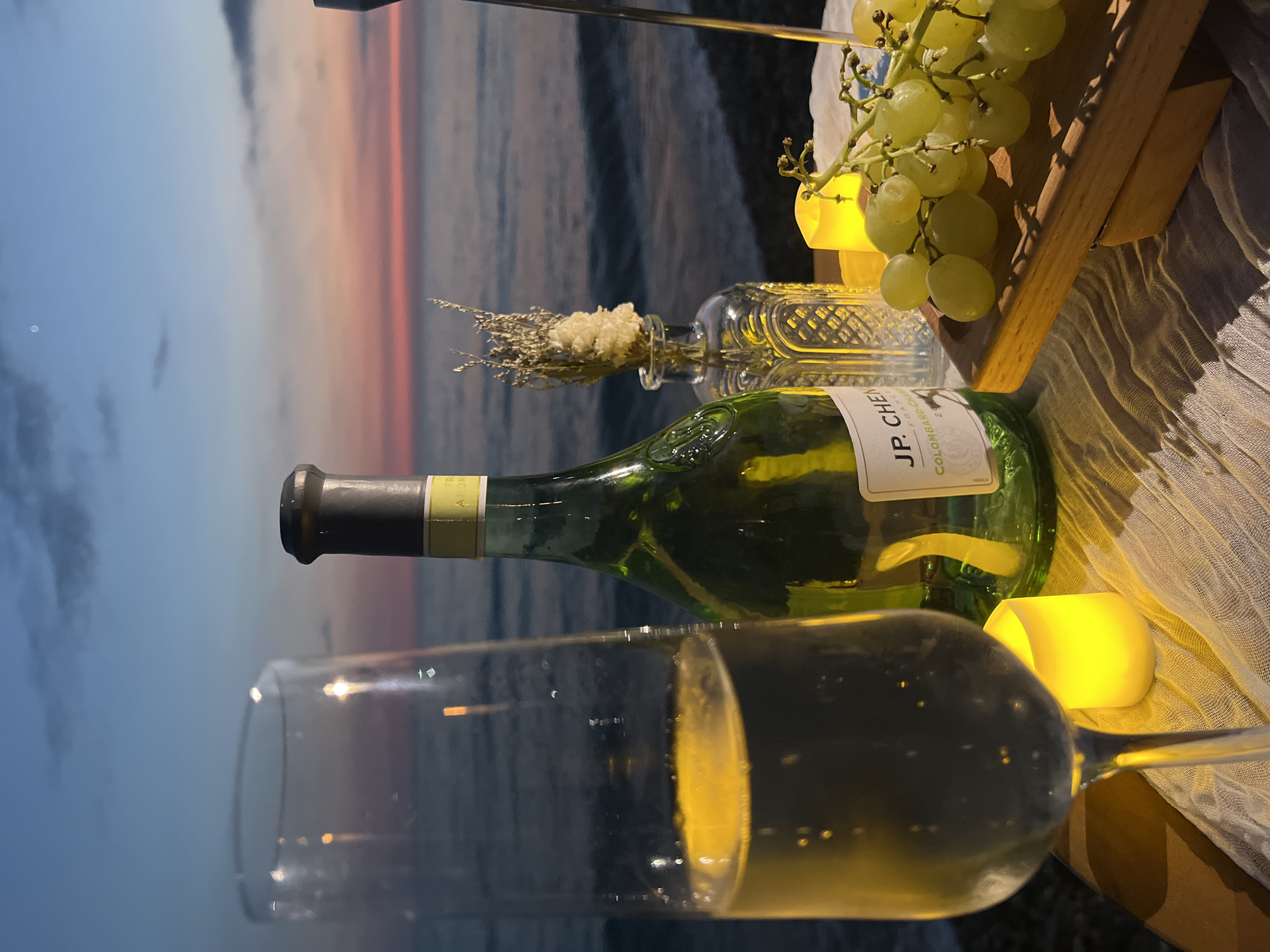 A table is set for a romantic evening on a beach in Sochi, Russia, at dusk. A flute of white wine, a bottle of JP. Chenet Colombard-Chardonnay, a small vase with dried flowers, two lit LED candles, and a bunch of green grapes on a wooden board are arranged on a white, gauzy cloth. The sea is visible in the background, with waves gently rolling onto the shore. The sky is a gradient of blue and pink, indicating the end of daylight. The scene evokes a sense of tranquility and celebration.