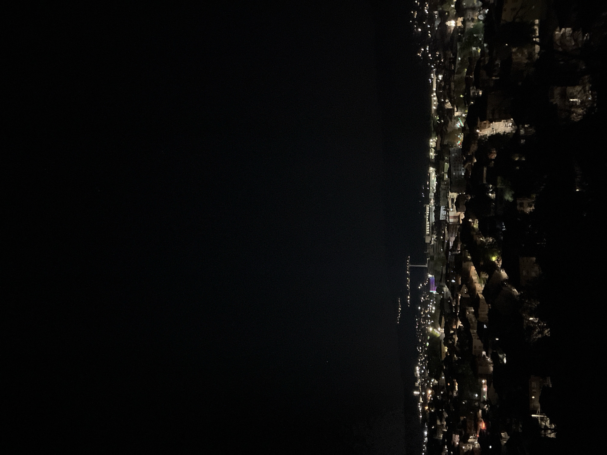 A night-time view in Sveti Konstantin, Bulgaria, shows a dark body of water bordered by the lit-up coastline. The coast is lined with buildings, illuminated trees, and streetlights. The lights create a dense, urban illumination pattern. No people are visible, only the urban environment.