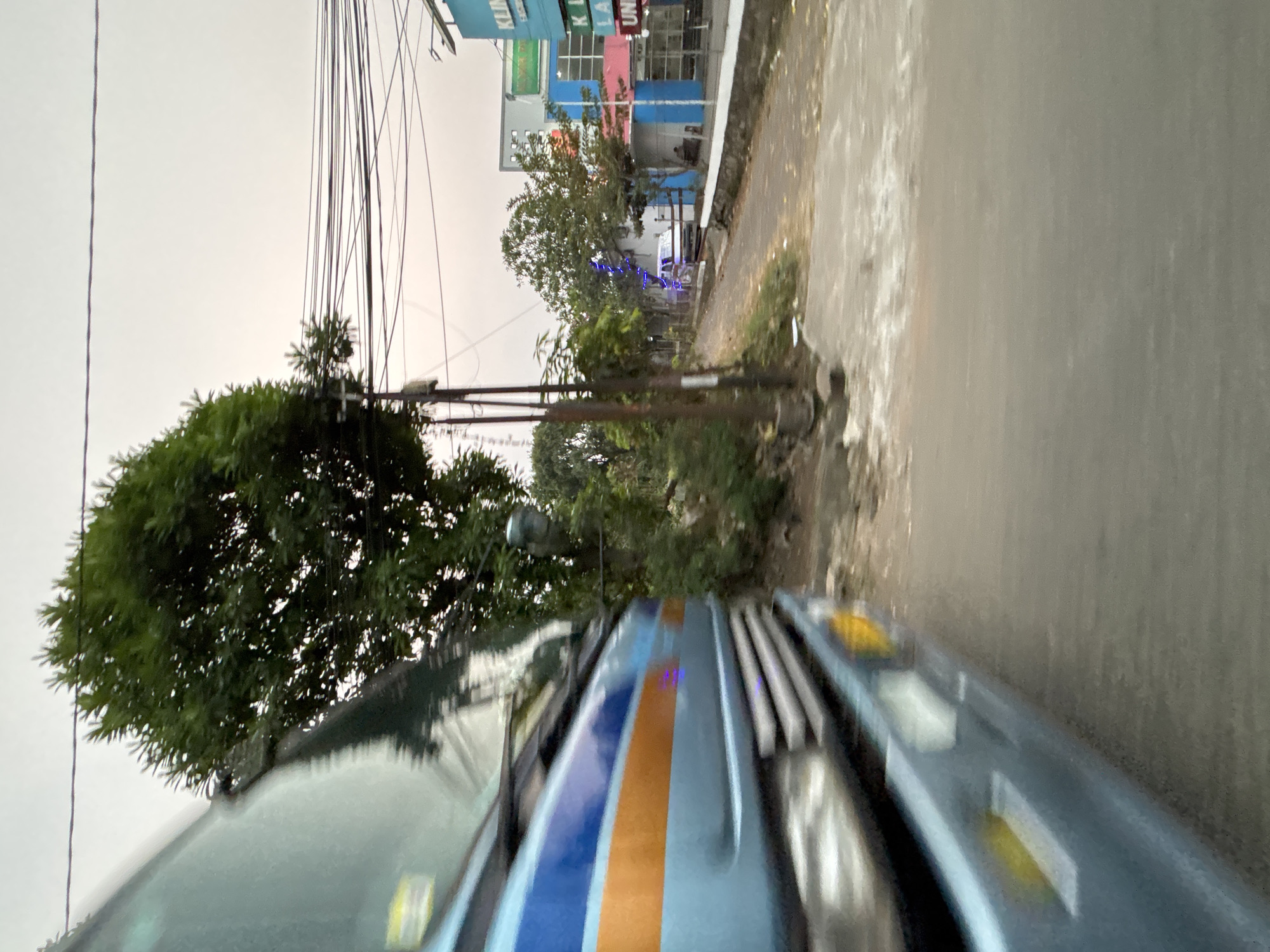 The image captures a view from a moving vehicle in Bekasi, Indonesia. In the foreground, the side and front of a blue and orange striped vehicle, possibly a bus or truck, are visible, exhibiting motion blur. To the right of the vehicle, a paved road curves slightly, bordered by a low stone wall. Beyond the road, several trees are present. Further in the background, a multi-story building with blue and pink accents is visible, featuring signage that reads "KLINIK" and "LA UN." The sky is overcast and light gray, suggesting a cloudy day. Electrical wires are strung across poles, typical of urban infrastructure. There are no clear human figures discernible in the image. The overall impression is a snapshot of a street scene in a developing urban area.