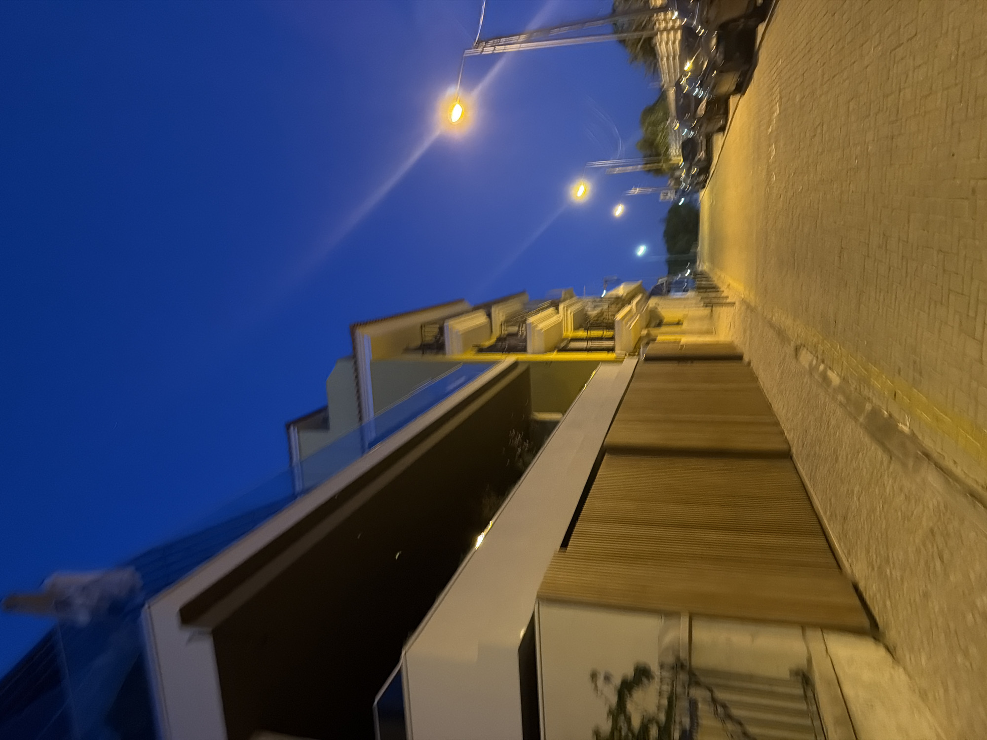 The image depicts a street in Cascais, Portugal at dusk. A modern building with a white facade and a dark balcony overhang occupies the left side of the frame. A wooden fence runs along the base of the building. The street is paved with interlocking bricks and illuminated by streetlights. Several cars are parked along the right side of the street. The sky is a deep blue, indicative of twilight.