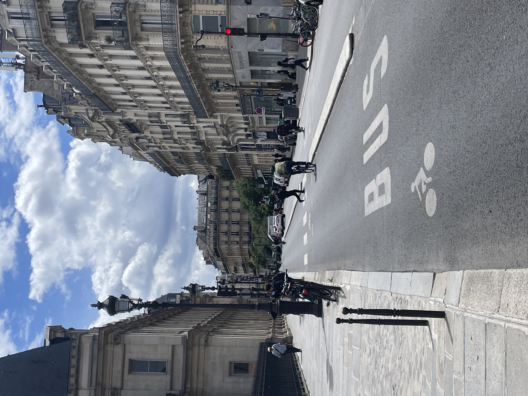The image shows a street scene in Paris, France, likely in a historic district, with ornate Haussmannian buildings lining a wide road. The buildings feature classical architectural elements, with balconies, mansard roofs, and large windows. The sky is partly cloudy, with dramatic white cumulus clouds against a blue backdrop.
On the left side of the street, a pedestrian walkway is paved with cobblestones and lined with ornamental black bollards. A man in a light-colored shirt and hat walks along the sidewalk, carrying a cane. A black metal fence runs parallel to the walkway. Lampposts with traditional designs are also visible along the street.
The road itself is asphalt, with lane markings and symbols. A large white "BUS" marking is painted on the road, along with a bicycle symbol indicating a dedicated bike lane or shared space. Several vehicles are present, including cars and what appears to be a white bus in the distance.
People are seen interacting with the urban environment. A cyclist in a green jacket is riding a bicycle in the foreground on the right side of the road. Another cyclist is visible further down the street. Pedestrians are crossing the street at a crosswalk, and others are walking along the sidewalk on the right. Bicycles are parked near the curb on the right side of the road. A traffic light is visible, showing a red signal. The scene conveys a sense of everyday urban life in Paris, with a mix of vehicular and pedestrian traffic, cycling infrastructure, and the characteristic architecture of the city.